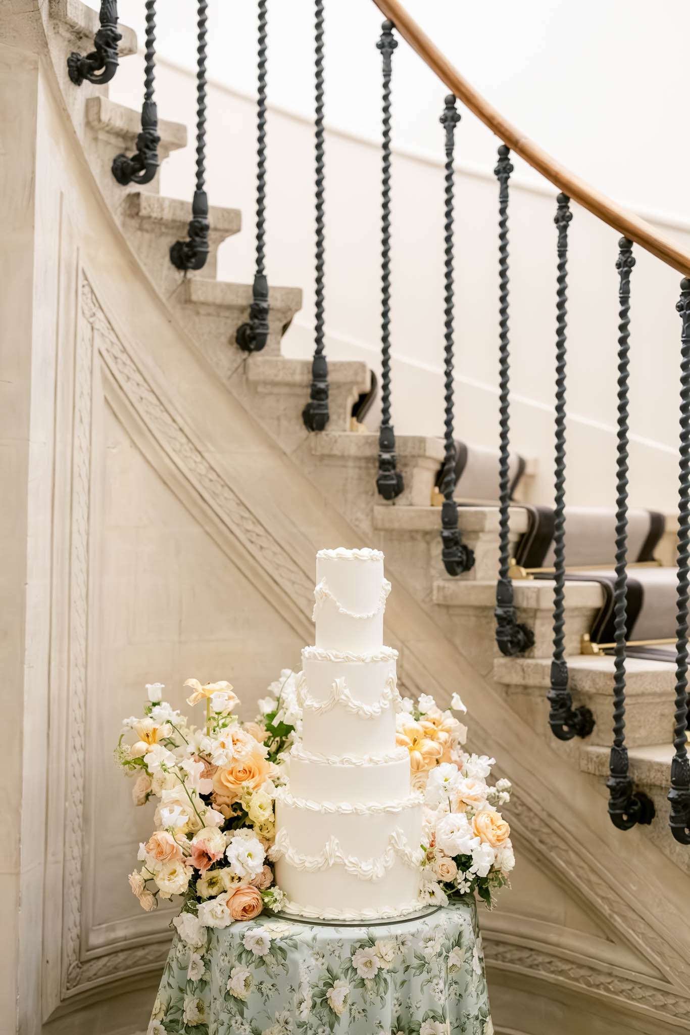 A five-tier white wedding cake with piped scroll and garland detailing is displayed on a small round table covered in a sage green floral-print linen, positioned at the base of a grand curved interior staircase. The cake is surrounded by lush floral arrangements featuring peach garden roses, white lisianthus, cream ranunculus, and pale yellow blooms. The staircase features ornate twisted wrought-iron balusters with a warm wood handrail and carved limestone or marble steps with decorative molding along the curved wall. The setting is a formal indoor venue with classic French architectural detailing. Medium shot, slightly angled to capture both the cake and the full sweep of the staircase. Potential venue feature image.