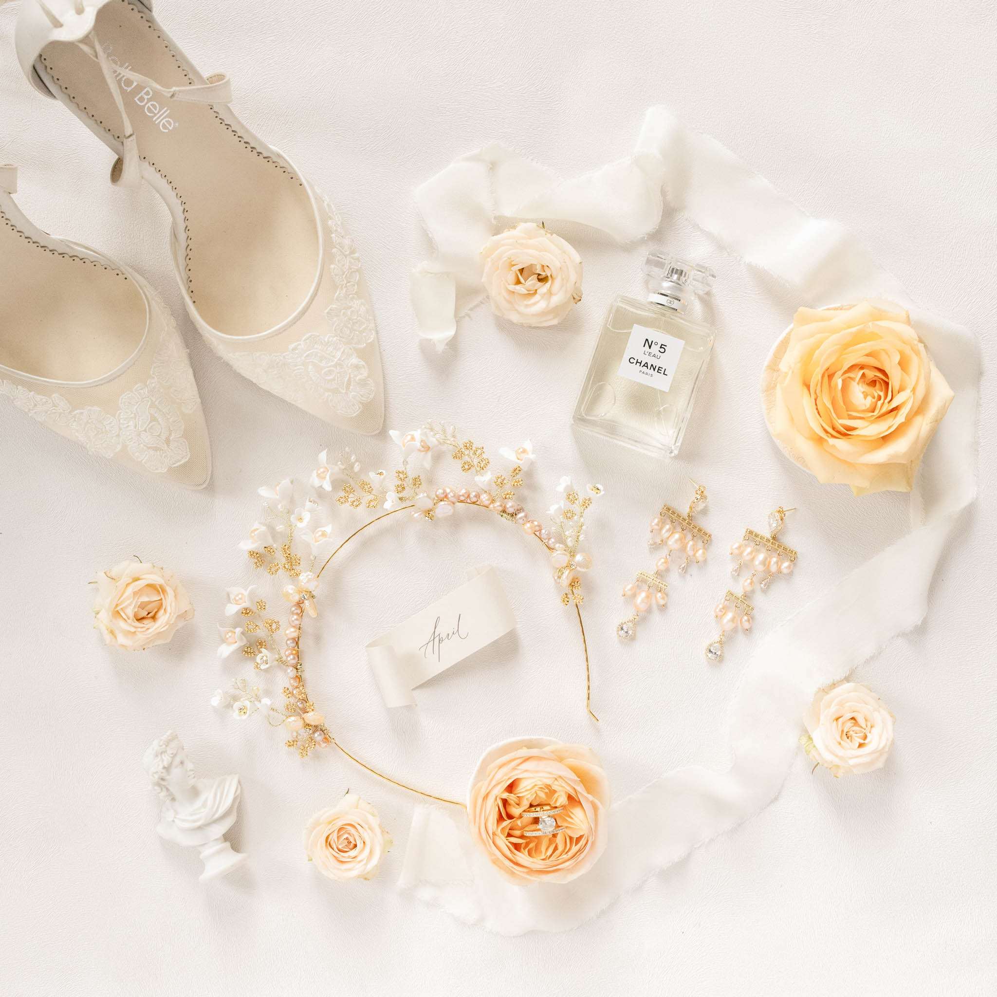 Bridal flat lay with Bella Belle lace heels, Chanel perfume, pearl headband, rings in a peach rose