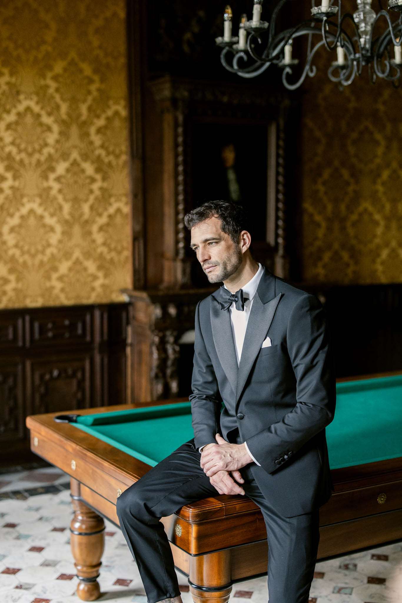 A groom portrait taken indoors in what appears to be a billiard room within a château or manor house. The groom is seated on the edge of a wooden-framed pool table with a green felt surface, wearing a charcoal tuxedo jacket with satin lapels, a white dress shirt, black bow tie, and a white pocket square. The room features gold damask-patterned wall covering, dark carved wood paneling, a wrought-iron candelabra chandelier with taper candles, and a patterned tile floor. The composition is a three-quarter portrait with natural light falling across the groom's face as he gazes to one side.