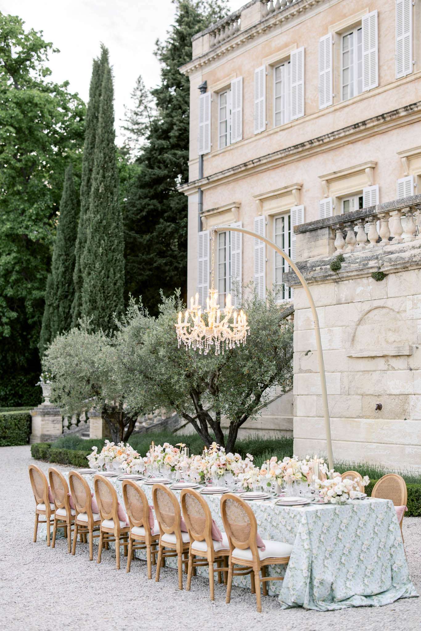 Long reception table with pastel floral arrangements and crystal chandelier on gold arc stand before French chateau