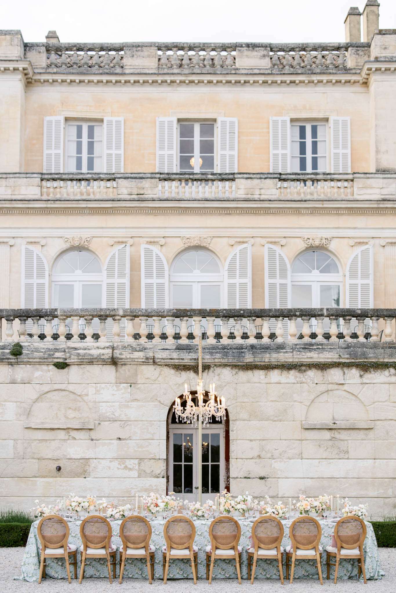 An outdoor reception table is set directly in front of a large French château, positioned on a gravel terrace at the base of the building's stone façade. The long rectangular table is dressed in a pale blue-grey floral-patterned tablecloth and lined with approximately ten natural oak cane-back Louis XVI-style chairs with cream upholstered seats. The tablescape features low arrangements of blush, peach, and ivory florals — likely ranunculus, roses, and sweet peas — with tall thin taper candles in glass holders running the length of the table. A crystal chandelier with lit candles is suspended on a tall gold stand above the center of the table, framed by an arched stone doorway behind it. The wide shot captures the full two-story château facade with white-shuttered arched and rectangular windows, a carved stone balustrade balcony, and classical French architectural detailing. Potential venue feature image.