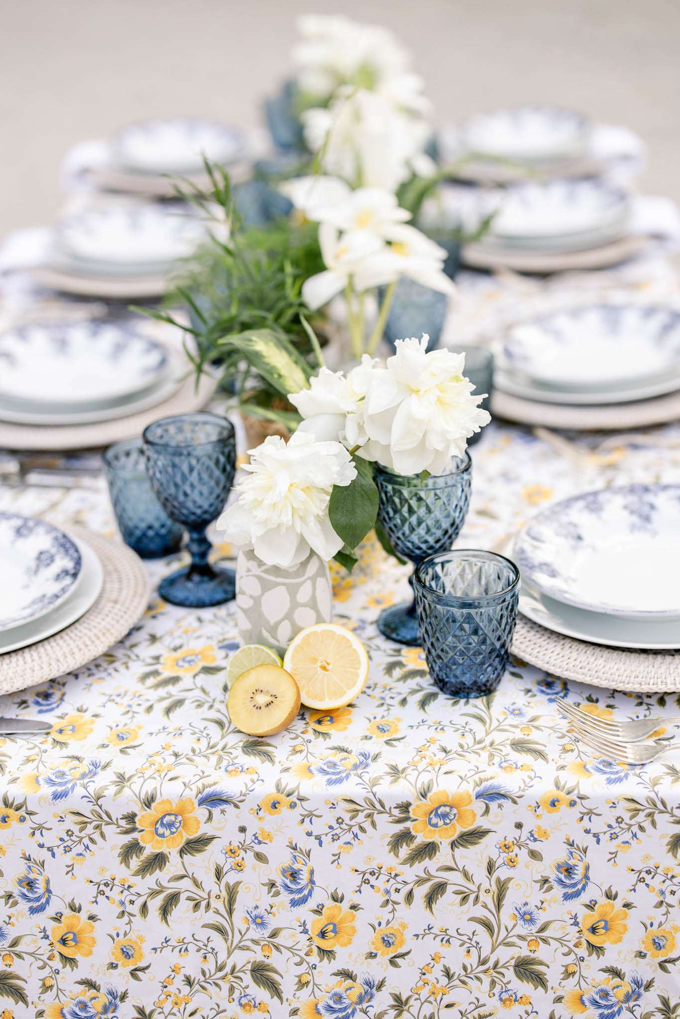 A close-up detail shot of a wedding reception tablescape styled with a blue and yellow floral-print tablecloth featuring botanical motifs on a white background. Place settings consist of white plates layered on woven rattan chargers, topped with blue-and-white patterned salad plates, paired with textured diamond-cut navy blue goblets and tumblers. The centerpiece runs along the table's length with white peonies, white ranunculus, and green foliage arranged in small vases, with halved lemons and a kiwi placed directly on the tablecloth as decorative elements. The overall color palette is navy blue, yellow, and white with a French country or Provençal styling influence.