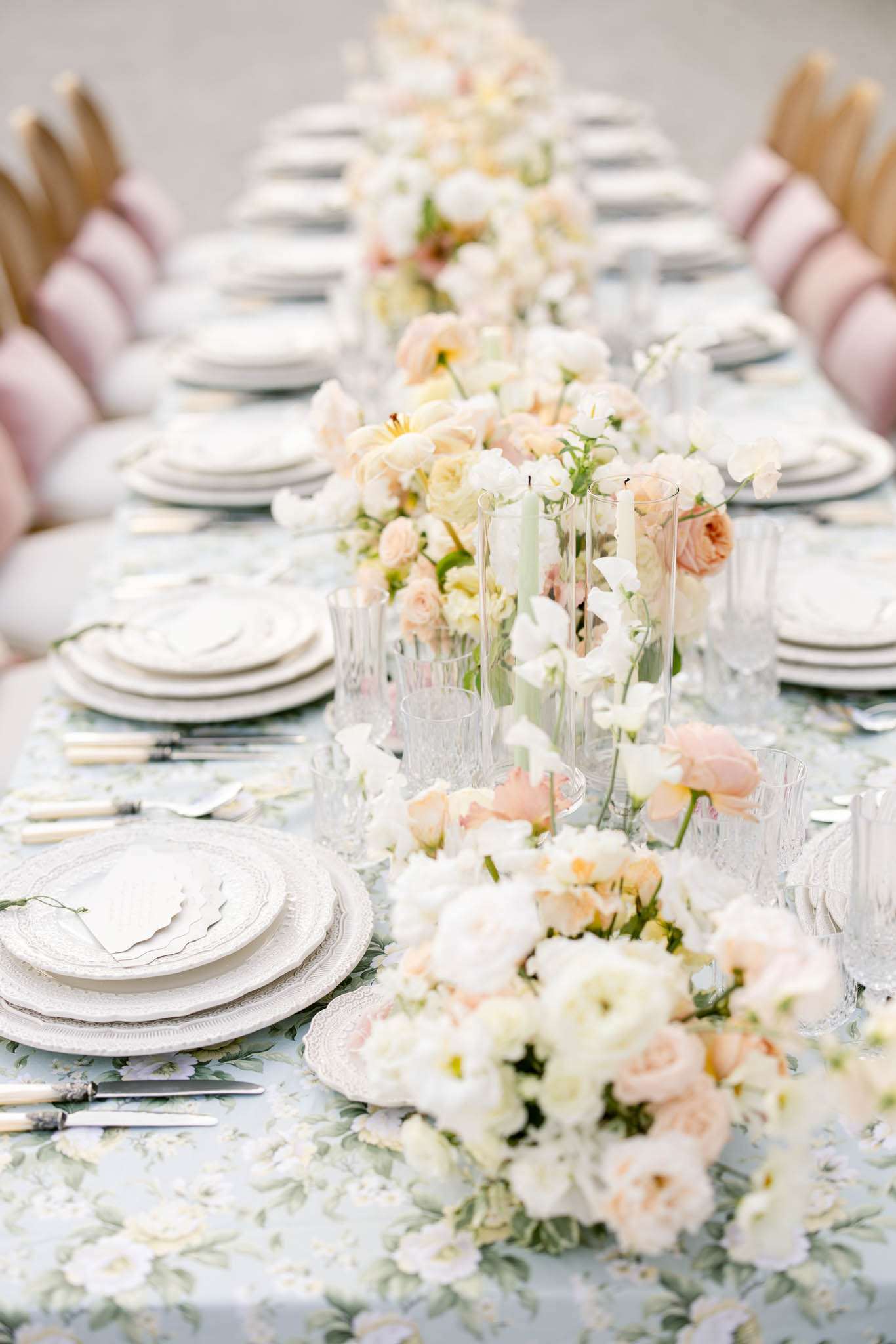 A close-up detail shot of a long wedding reception dining table set for approximately 16 guests, captured from a low angle looking down the length of the table. The tablecloth features a soft blue-grey floral print with sage green botanical motifs. Place settings consist of stacked white plates with scalloped and lace-edged chargers, cut-crystal glassware, and silver flatware with ivory handles. A continuous floral runner runs the full length of the table, composed of blush garden roses, cream ranunculus, white sweet peas, and pale yellow blooms arranged at low height with occasional slim sage green taper candles in glass holders interspersed throughout. Scalloped white menu cards are placed on top of each place setting. The chairs visible on both sides have dusty pink upholstered cushioned backs, creating a soft, romantic pastel palette overall. The styling is romantic and classic with a pastel color palette of blush, cream, and sage green.