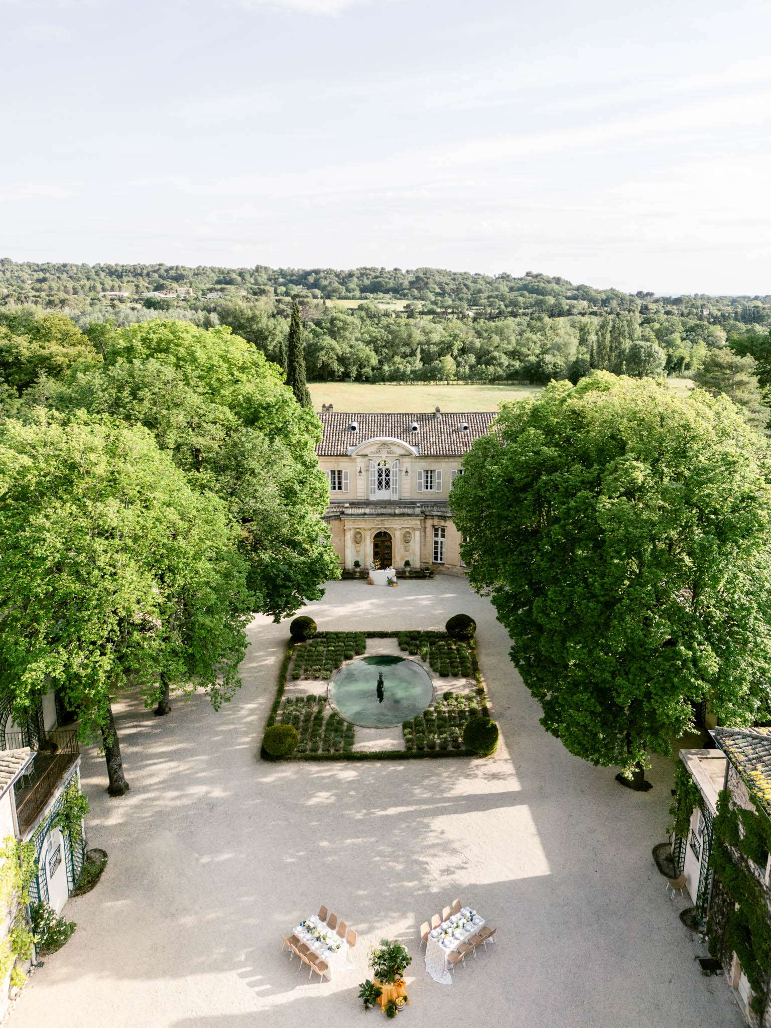 An aerial drone shot looking down the central axis of a classic French château property, with the stone manor house centered in the background featuring a terracotta tile roof, arched pediment, and ornate façade. The formal forecourt includes a circular fountain surrounded by a symmetrical parterre garden with low boxwood hedging and geometric plantings. Flanking the gravel courtyard are large mature trees forming a natural canopy on both sides, with outbuildings visible at the lower left and right edges. In the foreground, two rectangular reception dining tables are set with white linens, blush/nude chairs, and blue-toned floral centerpieces alongside potted topiaries, indicating outdoor reception setup. The overall styling is classic French formal garden with a refined, understated decor palette of white, blush, and blue. Potential venue feature image.