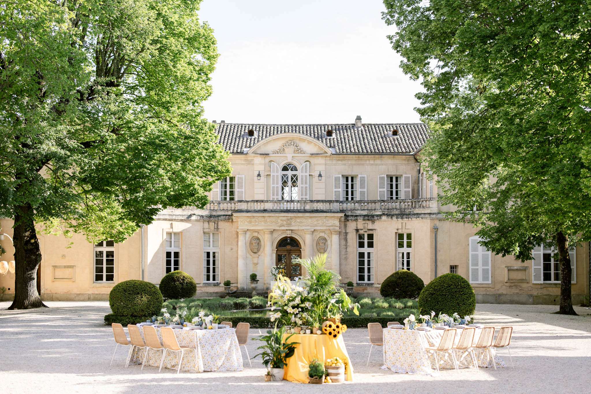 An outdoor reception setup photographed in a wide shot in the gravel courtyard of a classic French château with cream limestone façade, arched central entrance, white shuttered windows, and a tiled mansard roof. Several round dining tables are dressed in white and pale yellow floral-print tablecloths and surrounded by natural rattan and white chairs, arranged in a loose semicircle facing the building. A central display table is draped in a bright yellow cloth and decorated with sunflowers, tropical green foliage, potted plants, and small decorative items including what appear to be wooden barrels and citrus accents, creating a cheerful yellow and green color palette. The décor style is relaxed and garden-inspired, blending Provençal and botanical influences, with topiary box hedges and lavender beds visible in the formal garden between the tables and the château entrance. Potential venue feature image.