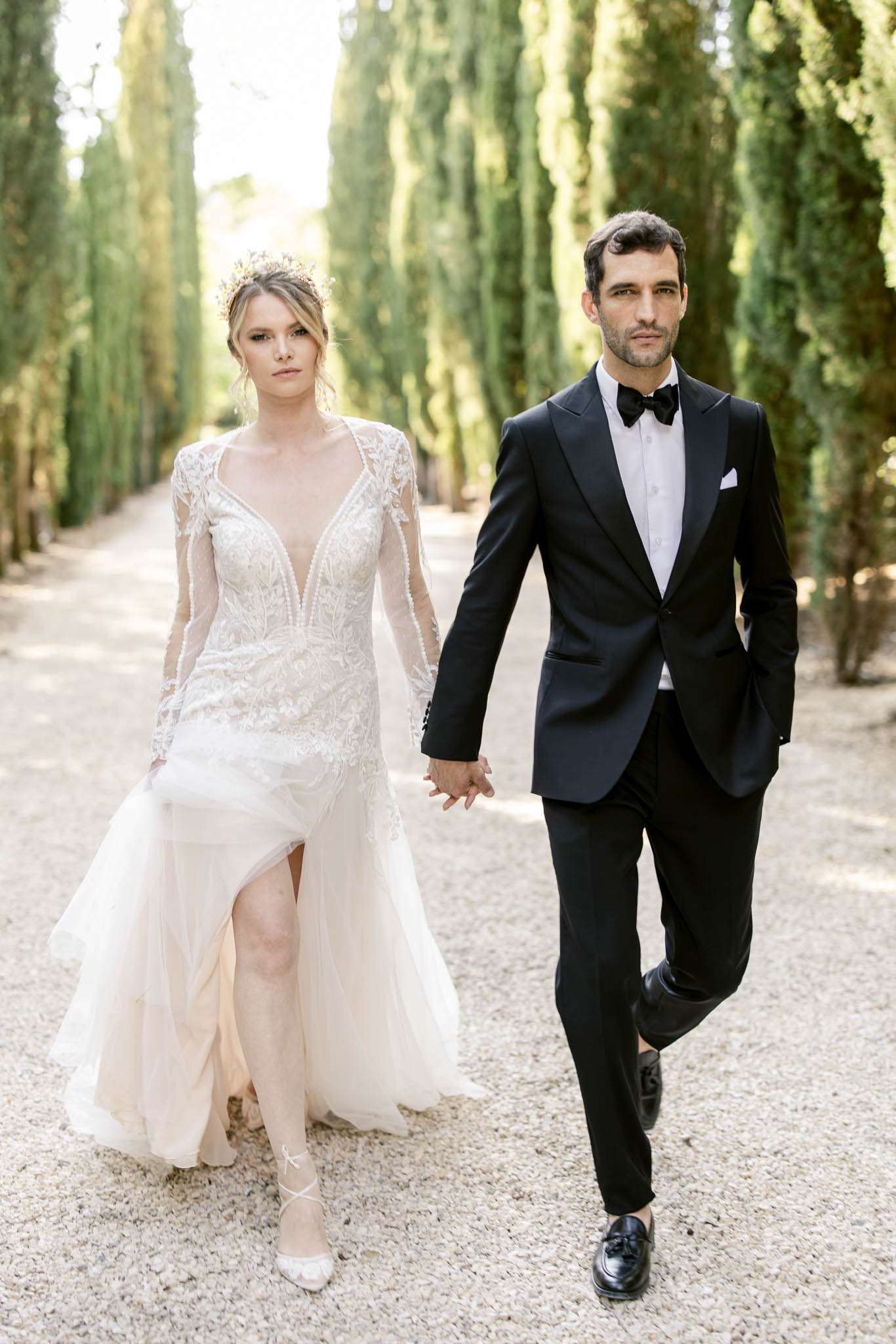 A couple portrait taken outdoors on a gravel path lined with tall cypress trees, shot as a mid-length portrait with the couple walking toward the camera while holding hands. The bride wears a long-sleeve, deep V-neck ivory lace gown with intricate embroidery across the bodice and sheer sleeves, paired with a flowing tulle skirt featuring a front slit, white strappy heels, and a gold floral crown headpiece. The groom wears a black tuxedo with satin lapels, a white dress shirt, black bow tie, white pocket square, and black loafers. The styling is modern-classic with a formal, editorial feel. The cypress tree alley creates a strong natural corridor framing the couple in the background.