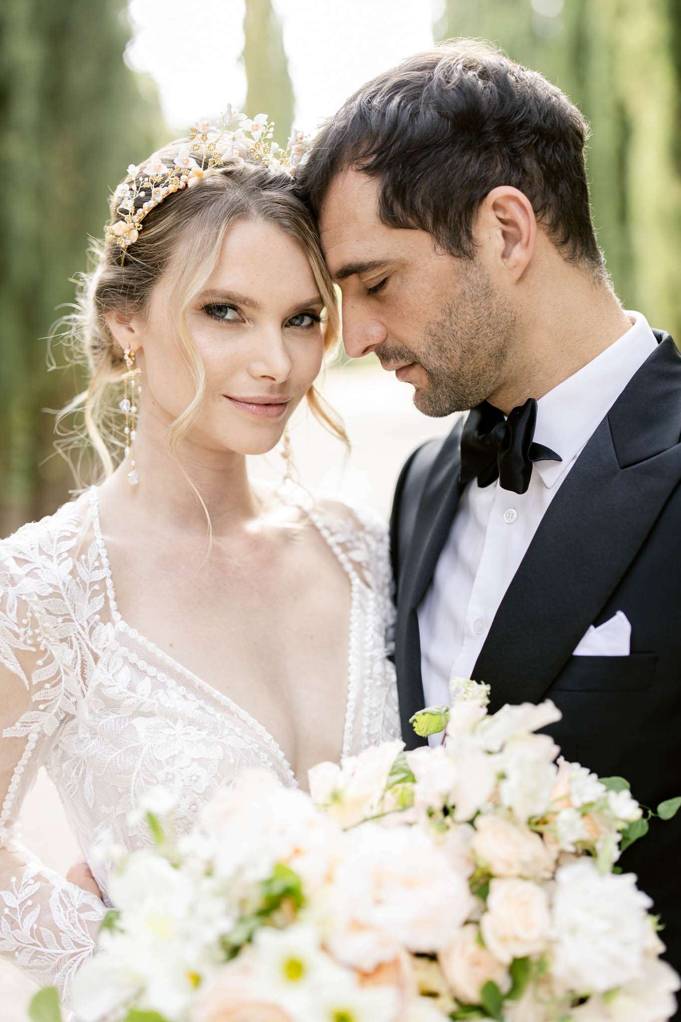 A close-up portrait of a bride and groom outdoors, with tall cypress trees softly blurred in the background. The bride wears a deep V-neck lace gown with floral embroidery and sheer long sleeves, paired with a gold and pearl floral headpiece and delicate drop earrings; she looks directly at the camera while the groom rests his head against hers with eyes downcast. The groom is dressed in a black tuxedo with a black bow tie and white pocket square. The bride holds a lush bouquet of white and blush peach roses with greenery, which fills the lower foreground of the frame. The overall styling is classic and polished with romantic details.