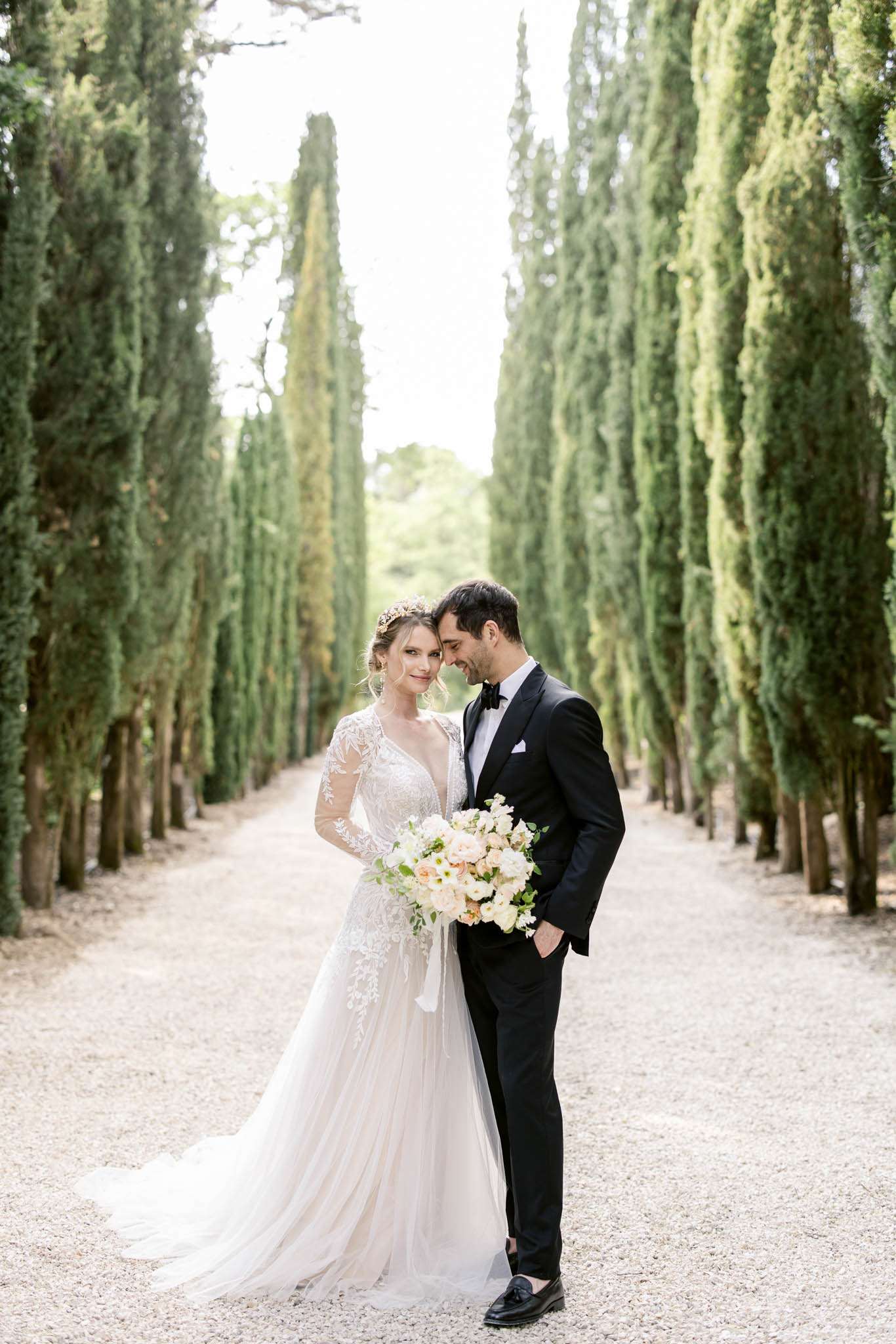 Bride in lace gown with blush bouquet and groom in tuxedo touching foreheads in cypress-lined allee