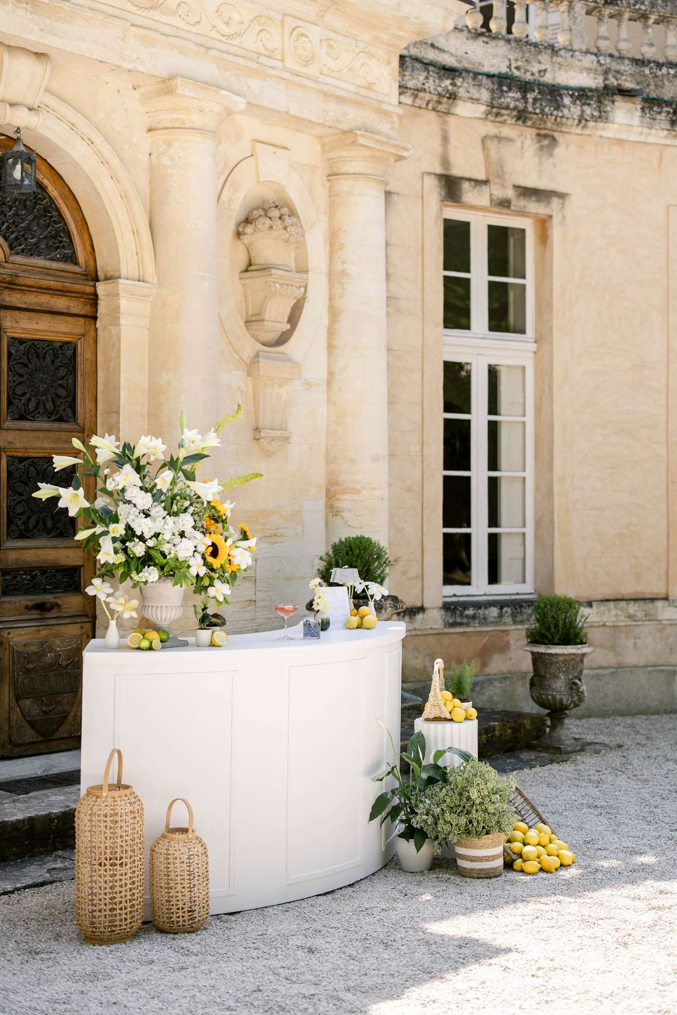 An outdoor cocktail hour bar setup positioned in front of a French château entrance, featuring a curved white bar counter topped with a large floral arrangement of white lilies, white stock flowers, and sunflowers in a stone urn, alongside scattered lemons and limes and a pink cocktail glass. The decor palette combines white, yellow, and green with a garden-fresh, classic French aesthetic. At the base of the bar, two wicker lanterns flank the front, while to the right a pedestal holds a basket of lemons, and potted herbs and greenery in a striped jute planter are arranged on the gravel courtyard floor. Additional loose lemons are grouped on the ground beside the display. The shot is a medium wide angle capturing both the full bar setup and the ornate carved limestone façade behind it. Potential venue feature image.
