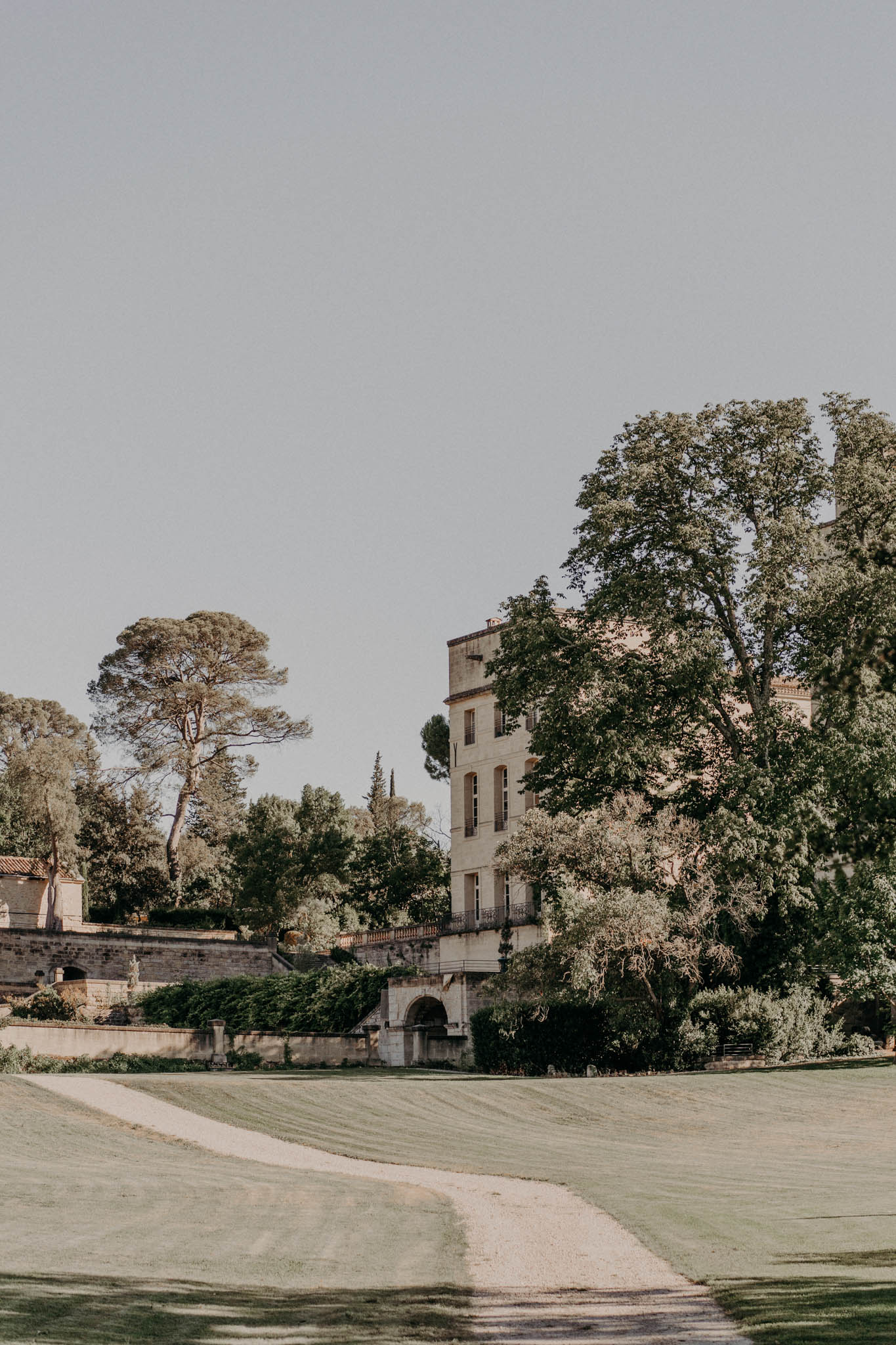 Cream stone chateau viewed across manicured lawn with gravel path, balustrades, and mature trees