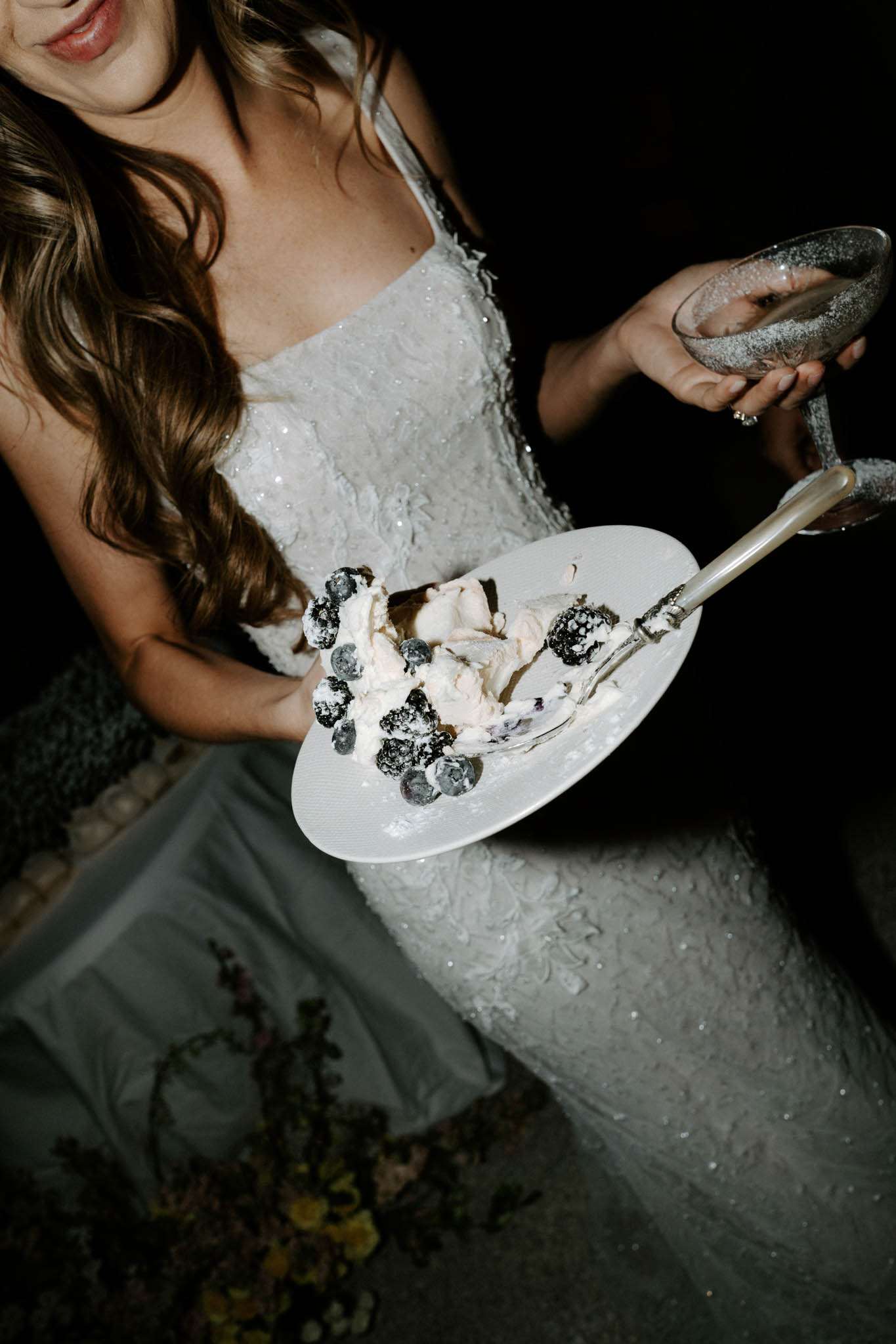 Close-up portrait of a bride during the reception, holding a white plate with a slice of white cake topped with fresh blueberries and a silver fork, alongside a champagne coupe glass. She is wearing a white textured sequined spaghetti-strap gown and has long wavy brown hair. The background is very dark, suggesting an evening event with limited ambient light. The shot is taken from slightly above, cropping out her face above the lips, and focuses on the cake plate and her dress details.