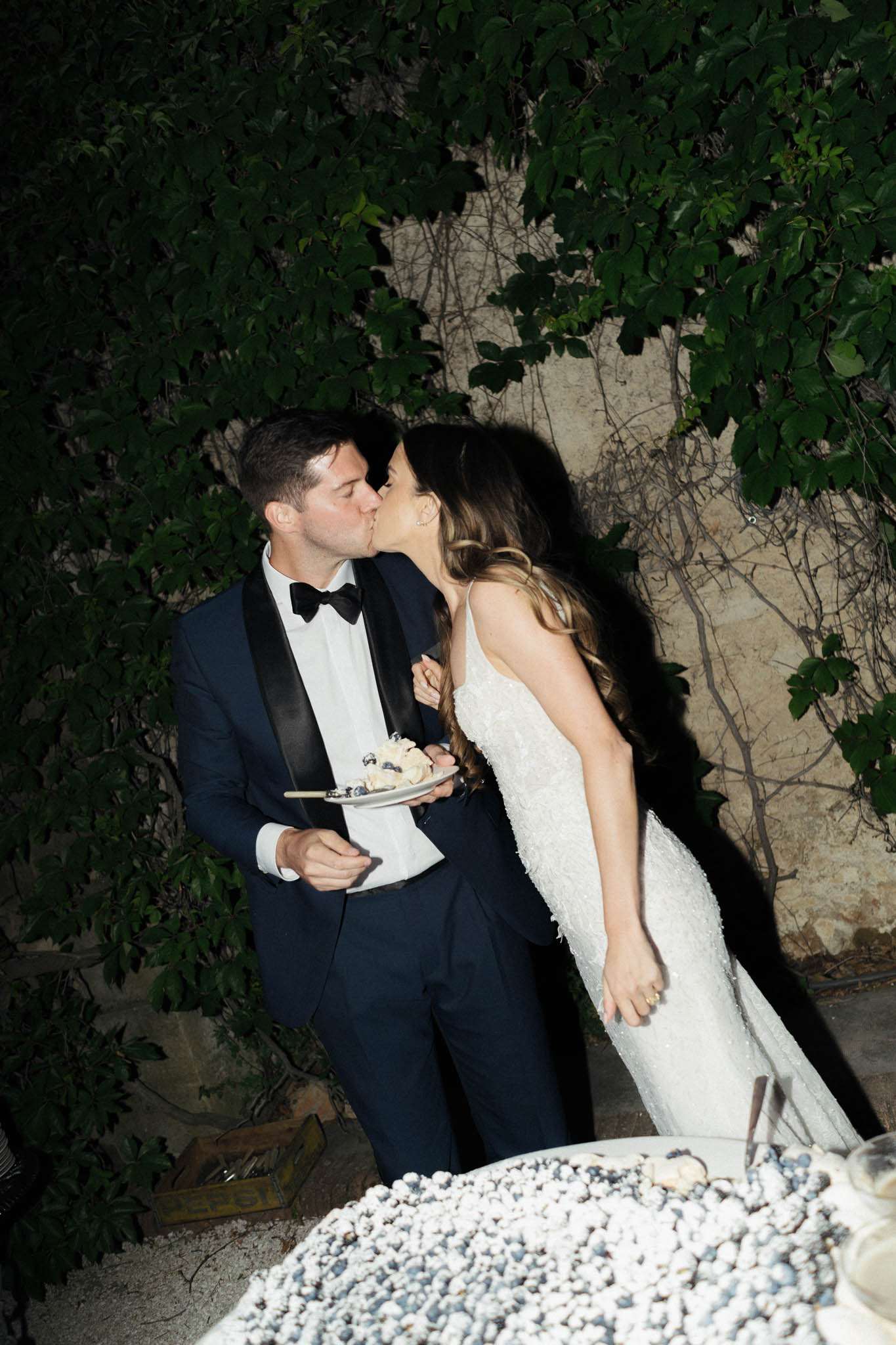 The bride and groom share a kiss immediately after cutting the cake during an outdoor evening reception. The groom is dressed in a navy tuxedo with black satin lapels and a black bow tie, and he holds a small plate with a slice of white cake. The bride wears a fitted, spaghetti-strap white gown with allover beaded or sequined embellishment and has long wavy hair. In the foreground, the wedding cake is partially visible — it appears to be a white cake decorated with what look like blueberries or dark berries. The shot is taken from a slightly elevated angle, framing the couple against a stone wall backdrop with climbing vines. The lighting is low and dramatic, characteristic of nighttime outdoor reception photography.