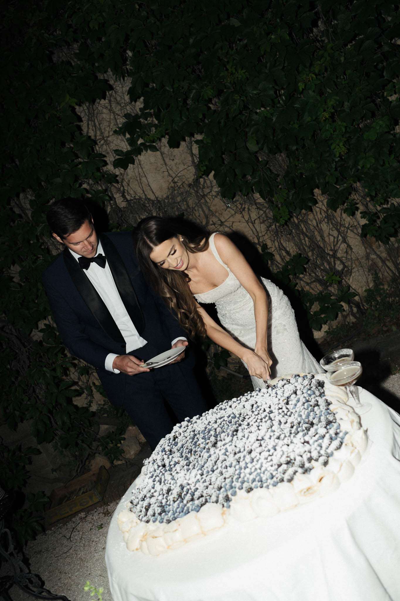 The bride and groom are cutting their wedding cake during an outdoor evening reception, photographed from a slightly elevated angle. The groom wears a black tuxedo with a black bow tie and holds a plate, while the bride wears a fitted, textured ivory gown with a low back and long wavy brunette hair. The wedding cake is a large, round white cake densely topped with fresh blueberries dusted with powdered sugar and decorated with cream swirls along the base. The setting is outdoors at night against a stone wall covered in climbing vines, with the scene lit by a warm spotlight. Two champagne flutes are visible on the cake table, which is draped in a white cloth.