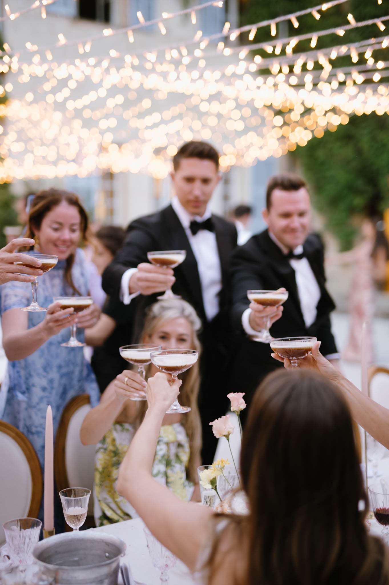 An outdoor evening reception toast captured mid-celebration, with approximately six guests raising coupe glasses filled with what appear to be espresso martinis toward the camera. Two men in black tuxedos with bow ties stand at the back, while a woman in a floral yellow-and-white dress and another in a blue floral dress participate in the toast. The foreground shows the back of a guest seated at a reception table dressed in a white outfit, with small bud vases holding blush pink and pale yellow flowers, a champagne flute, crystal glassware, a pale pink taper candle, and a wine cooler visible on the table. Dense warm-toned fairy lights are strung overhead in a canopy formation, creating a golden bokeh effect across the upper portion of the frame. The shot is taken from a low angle at table level, giving a dynamic upward perspective toward the raised glasses.
