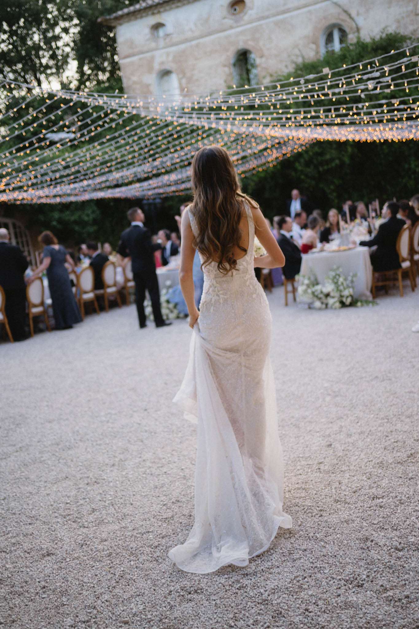 The bride is photographed from behind as she walks toward an outdoor evening reception set in the courtyard of a French stone château. She wears a fitted, lace-detailed ivory gown with a low open back and a trailing skirt, and her long wavy dark hair falls loose down her back. Overhead, dense rows of warm fairy lights are strung in a canopy formation across the courtyard, casting a golden glow over the scene below. Guests are seated at round tables dressed in white linens with white floral arrangements and gold chiavari chairs, while additional guests mingle standing nearby. The composition is a medium full-length shot taken from ground level, with the bride sharp in the foreground and the reception tables softly blurred in the background.