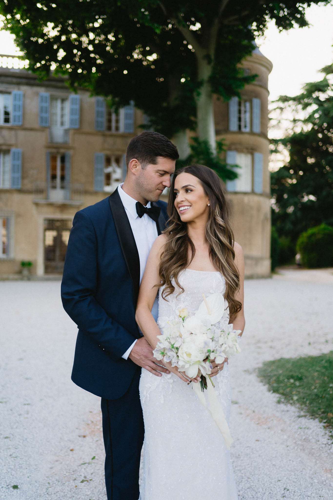 Bride in strapless beaded gown with white peony bouquet and groom in navy tuxedo in front of a French chateau