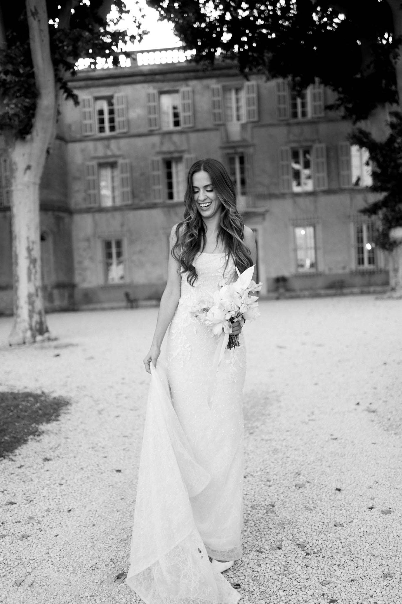 This black-and-white bridal portrait shows a bride walking across a gravel courtyard in front of a large French château, photographed at dusk with interior lights visible through the ground-floor windows. The bride wears a strapless lace A-line gown with a flowing train, which she lifts slightly with one hand, and carries a loose, unstructured bouquet of light-toned blooms with large petals — likely tulips or ranunculus. She has long, wavy dark hair worn down and is laughing with her eyes cast downward. The image is shot in high-contrast black and white, with bright highlights on the dress and soft mid-tones throughout the background architecture. The composition is a full-length portrait with the château façade centered behind her.