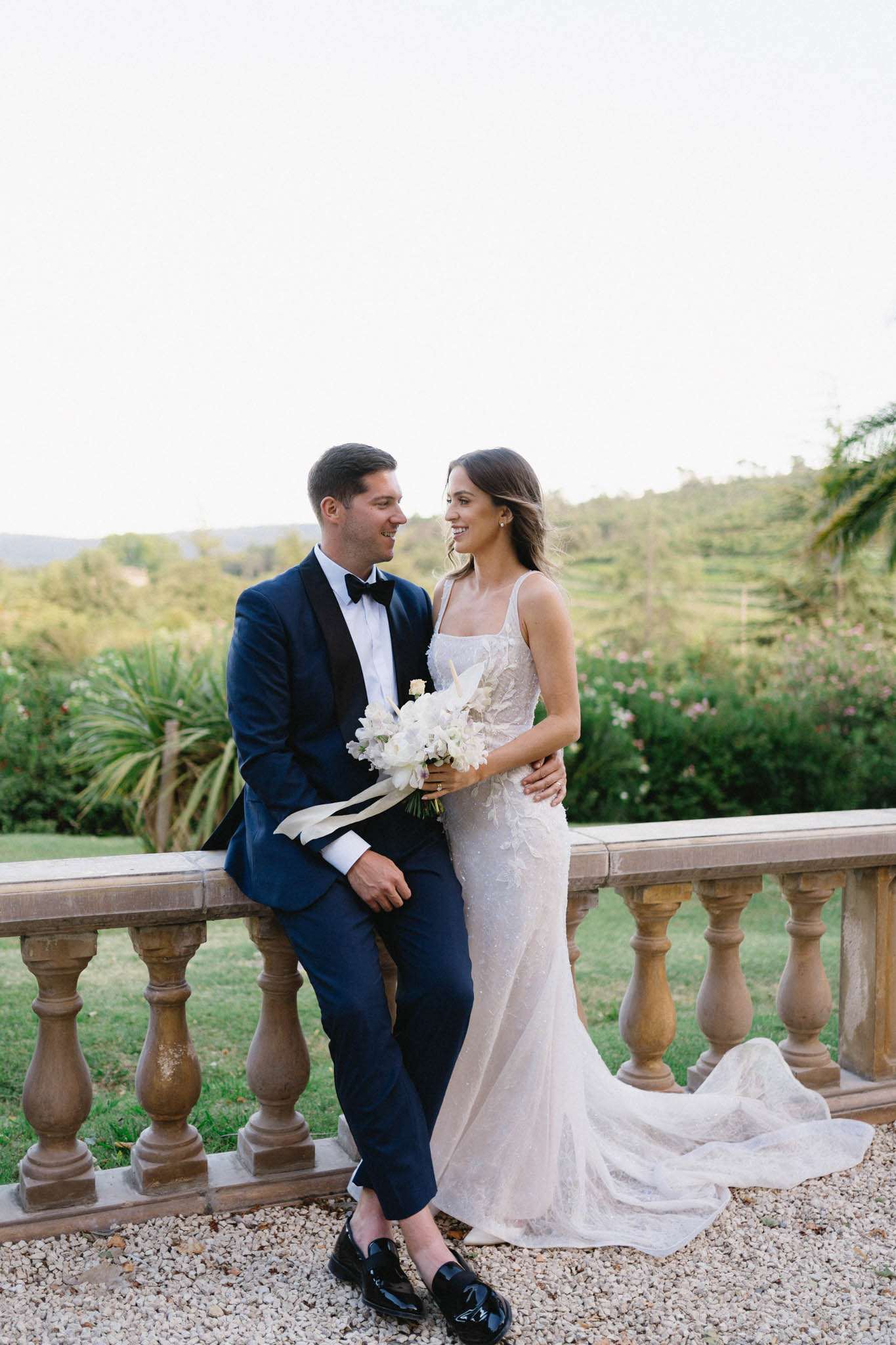 Groom on balustrade in navy tuxedo beside bride in beaded ivory gown with peony bouquet at garden terrace