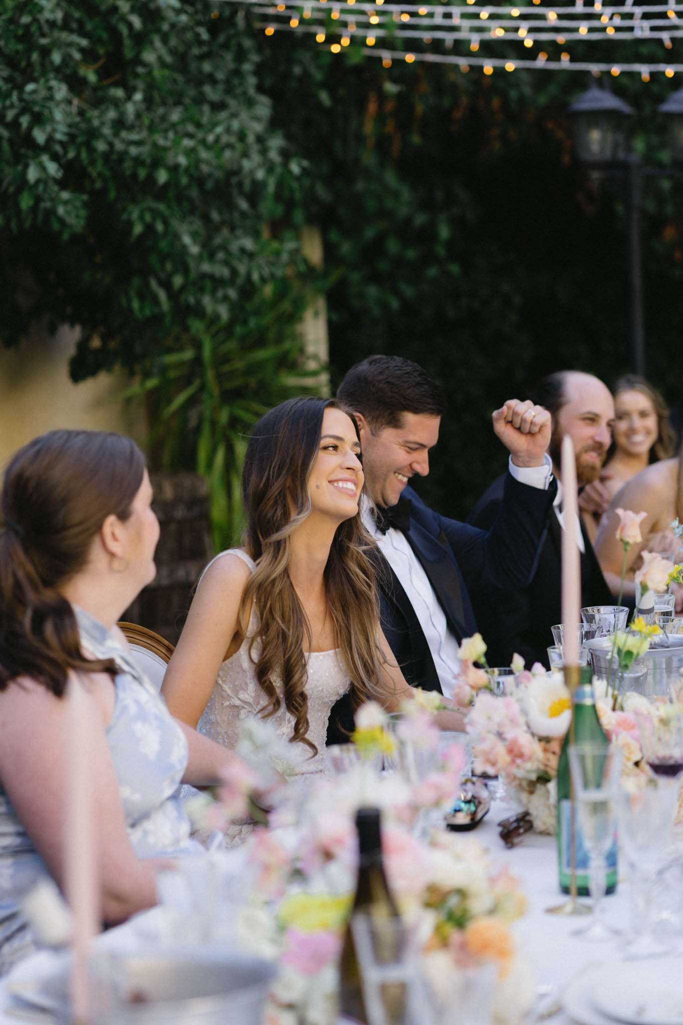 Couple laughing at reception table with blush and peach blooms, gold candle holders, under string lights