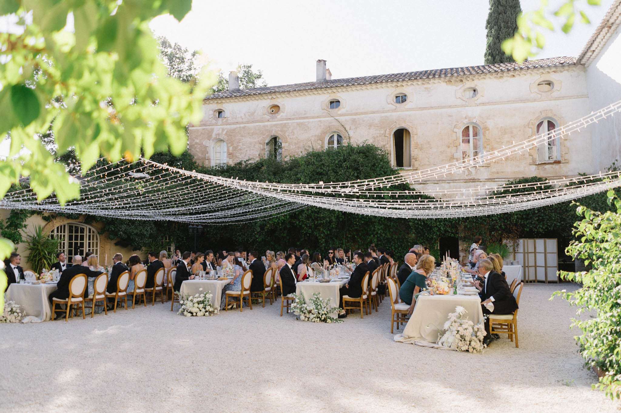 Elevated view of outdoor courtyard reception with U-shaped banquet tables, crystal canopy installation, and Provencal ston...