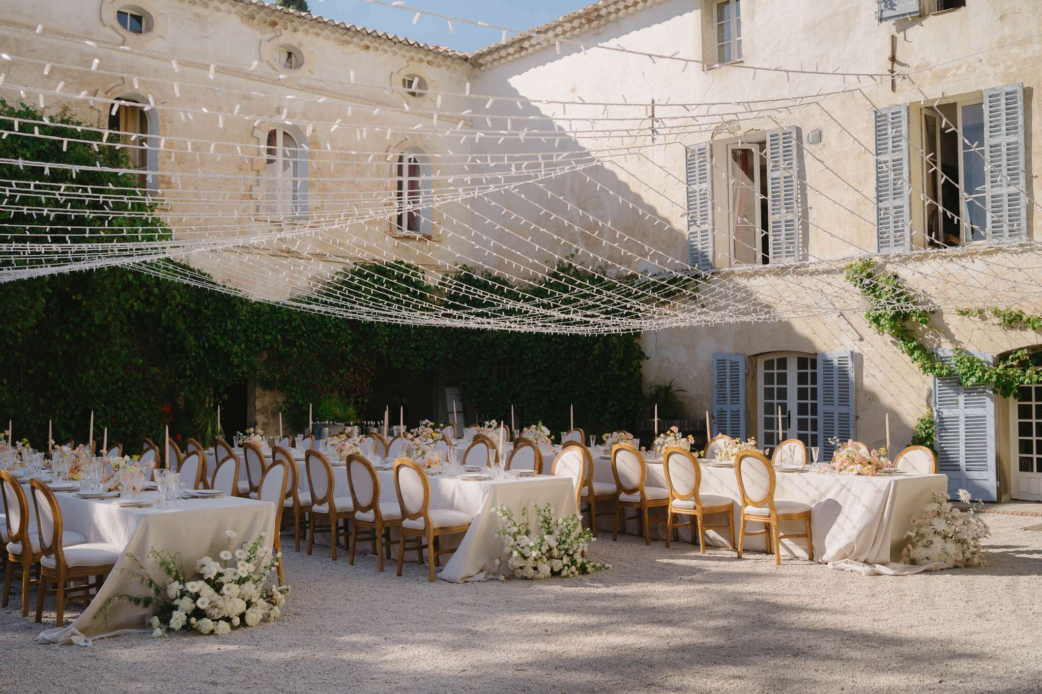 An outdoor wedding reception setup in the courtyard of a French château, with long rectangular tables dressed in ivory linen tablecloths arranged in parallel rows. The tables are lined with gold-framed Louis XVI-style chairs with ivory upholstered backs and seats. Table decor includes clusters of blush, cream, and white florals — appearing to include ranunculus, garden roses, and daisies — along with tall taper candles in blush and nude tones. Low floral arrangements of cream and white blooms are placed on the gravel ground at the base of the tables. Overhead, an elaborate canopy of criss-crossing strings hung with what appear to be white paper or fabric ribbon elements stretches across the entire courtyard. The wide-angle shot captures the full reception layout set against the château's stone facade with grey-blue shuttered windows and ivy-covered walls. Potential venue feature image.