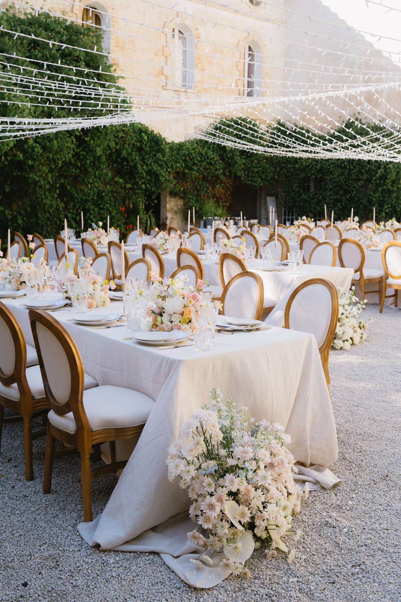 An outdoor wedding reception setup in the courtyard of a French chateau, featuring multiple long banquet tables dressed in ivory linen tablecloths that pool slightly onto the gravel ground. The tables are lined with gold-framed Louis XVI-style chairs with cream upholstered seats and backs. Centerpieces consist of low arrangements of blush pink, peach, soft yellow, and white flowers including dahlias, ranunculus, and delicate filler blooms, paired with tall taper candles in soft peach tones. A large floor-level floral arrangement of white daisies, blush chrysanthemums, and pale blue accent blooms sits at the base of the nearest table end. Overhead, white fairy light strings are draped in a canopy pattern across the courtyard. Crystal glassware and white china place settings complete each setting. The wide shot shows the full reception layout set for a large number of guests, with the honey-colored stone facade of the chateau and dense ivy-covered walls visible in the background. Potential venue feature image.