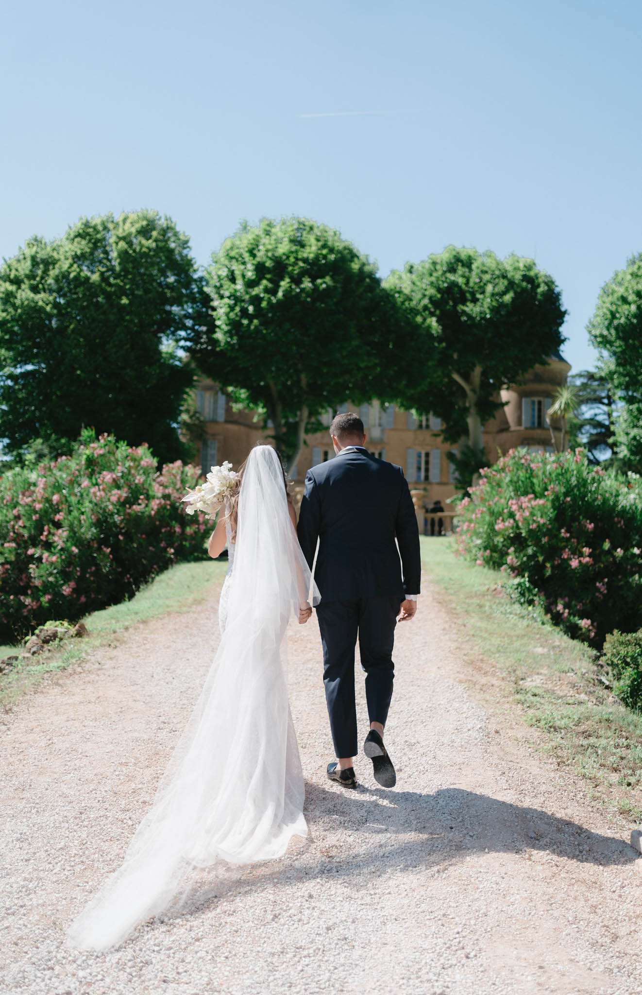 A couple portrait shot from behind, showing the bride and groom walking hand-in-hand along a gravel driveway toward a French château in the background. The bride wears a fitted white gown with a long cathedral-length veil trailing on the ground, and carries a bouquet of white and cream blooms. The groom wears a navy suit with black loafers. The driveway is lined with manicured round-canopied trees and pink-flowering shrubs, with the stone façade of the château visible at the end of the path. The composition is a wide portrait shot with strong central symmetry, taken in bright midday light. Potential venue feature image.