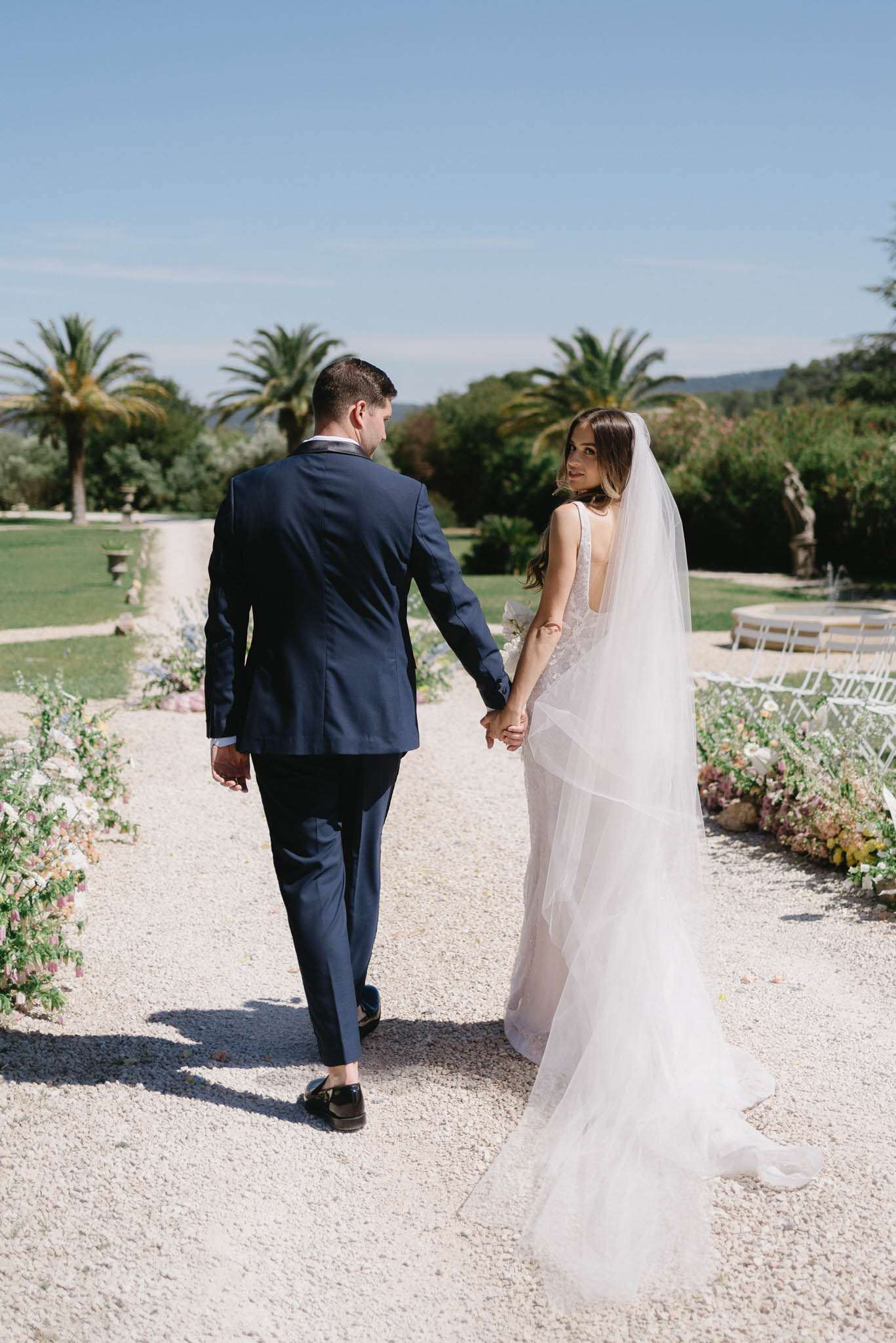 Bride and groom walking down gravel path lined with flowers, bride looking back showing lace gown open back and veil