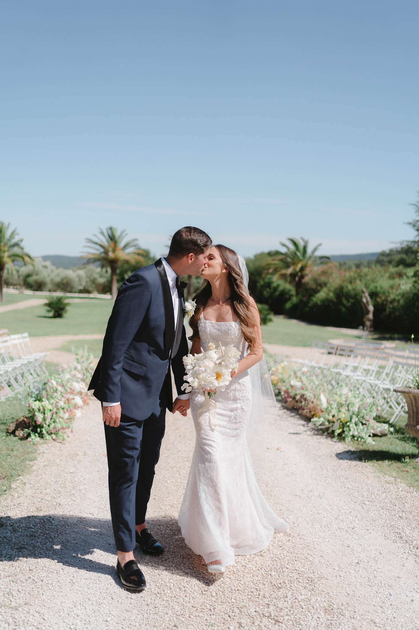 Bride and groom kiss walking along outdoor aisle flanked by white chairs and pink and white floral arrangements