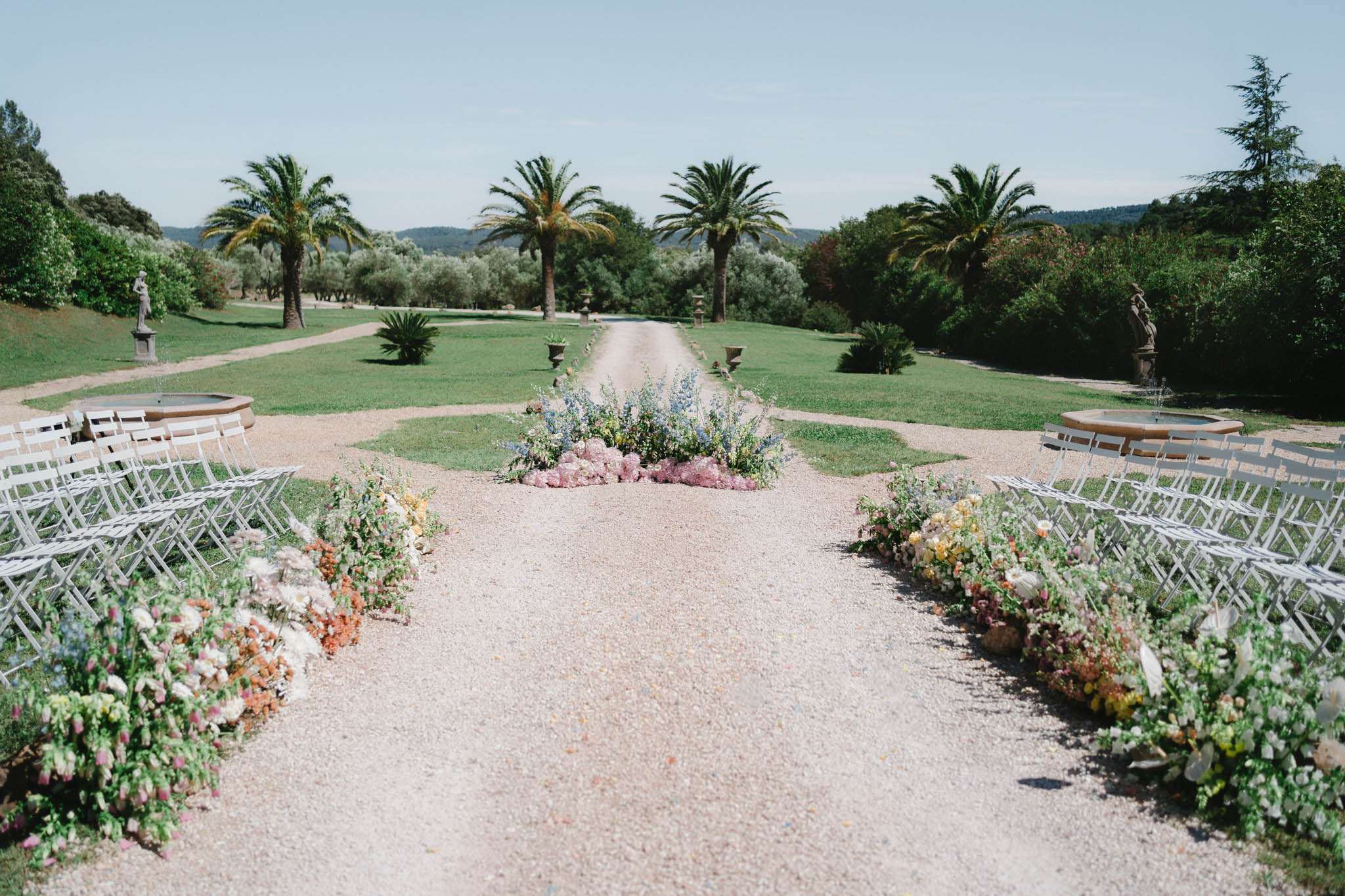 Ceremony aisle with meadow-style florals stone fountains statuary and palm-lined allee at estate
