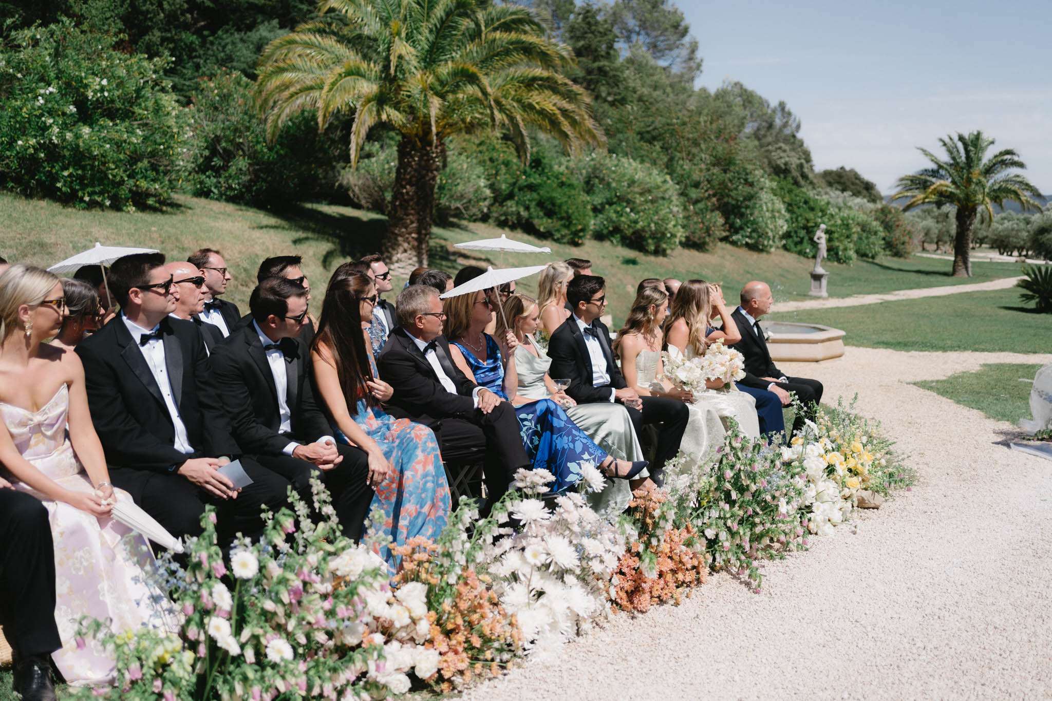 Wedding guests seated on benches along a gravel aisle lined with cream and peach floral arrangements in a formal garden