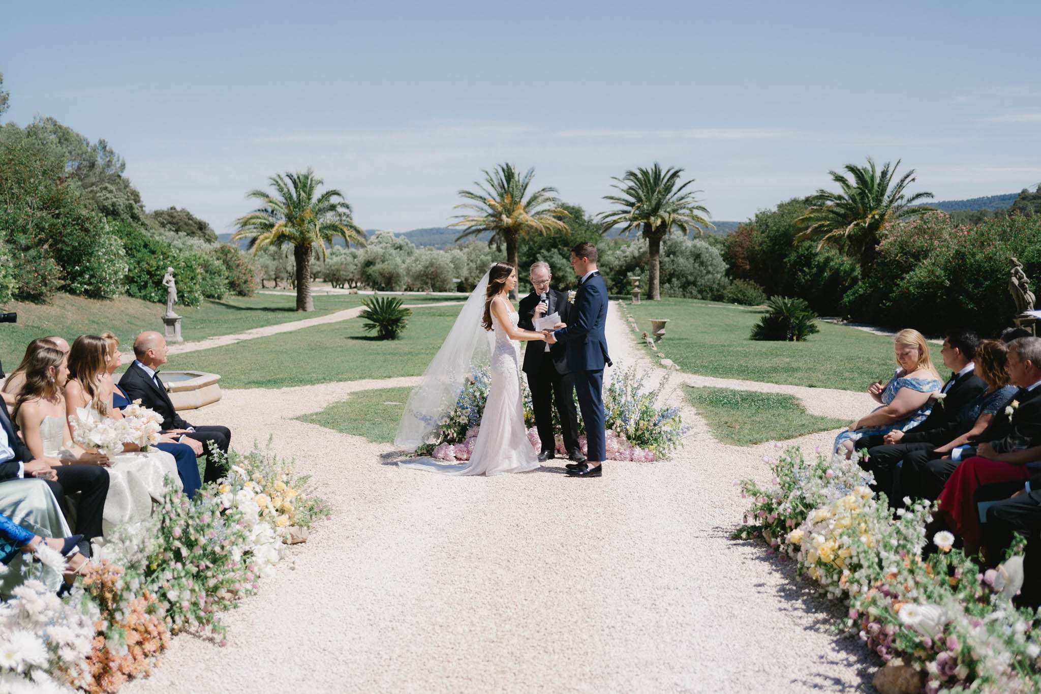 Couple at altar on gravel aisle with pastel wildflower arrangements and Provencal estate fountain backdrop