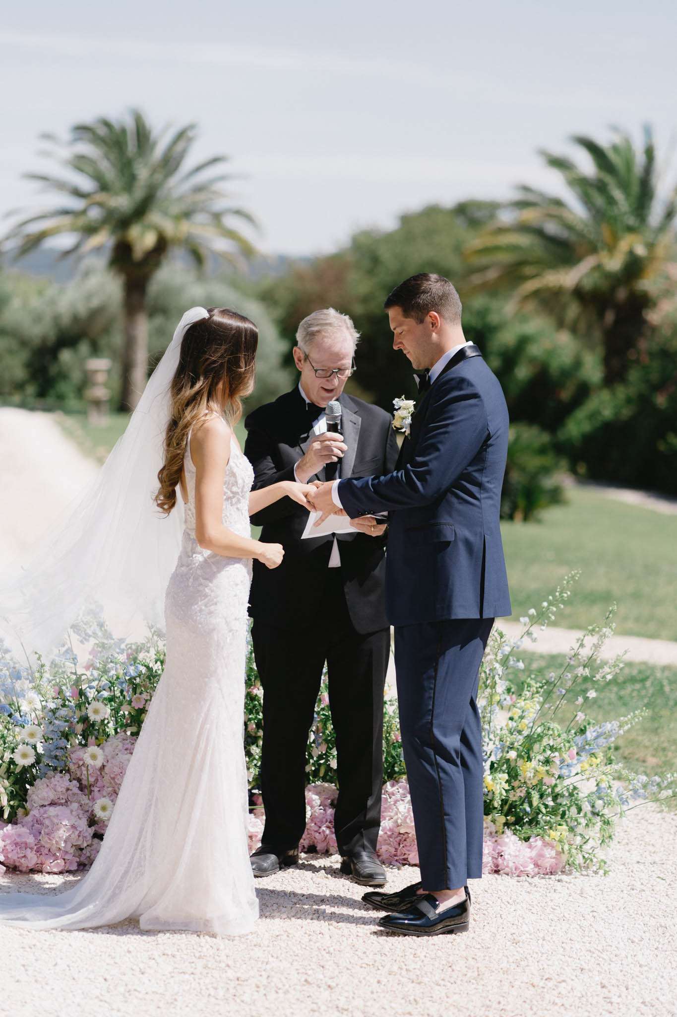 An outdoor wedding ceremony ring exchange taking place on a gravel path, with the groom placing a ring on the bride's finger while the officiant reads from papers and holds a microphone. The bride wears a fitted white lace gown with a long cathedral-length veil, and the groom is dressed in a navy blue suit with black lapels and black loafers, wearing a white and cream boutonniere. The officiant is in a dark suit. Low floral arrangements line the ceremony aisle on both sides, composed of blush pink hydrangeas, white daisies, pale blue delphiniums, and soft yellow blooms with trailing greenery, creating a garden-style, soft pastel palette. The setting is an outdoor estate with a manicured lawn and tall palm trees in the background, suggesting a South of France or Mediterranean property. The composition is a medium full-length portrait shot taken in bright midday light.