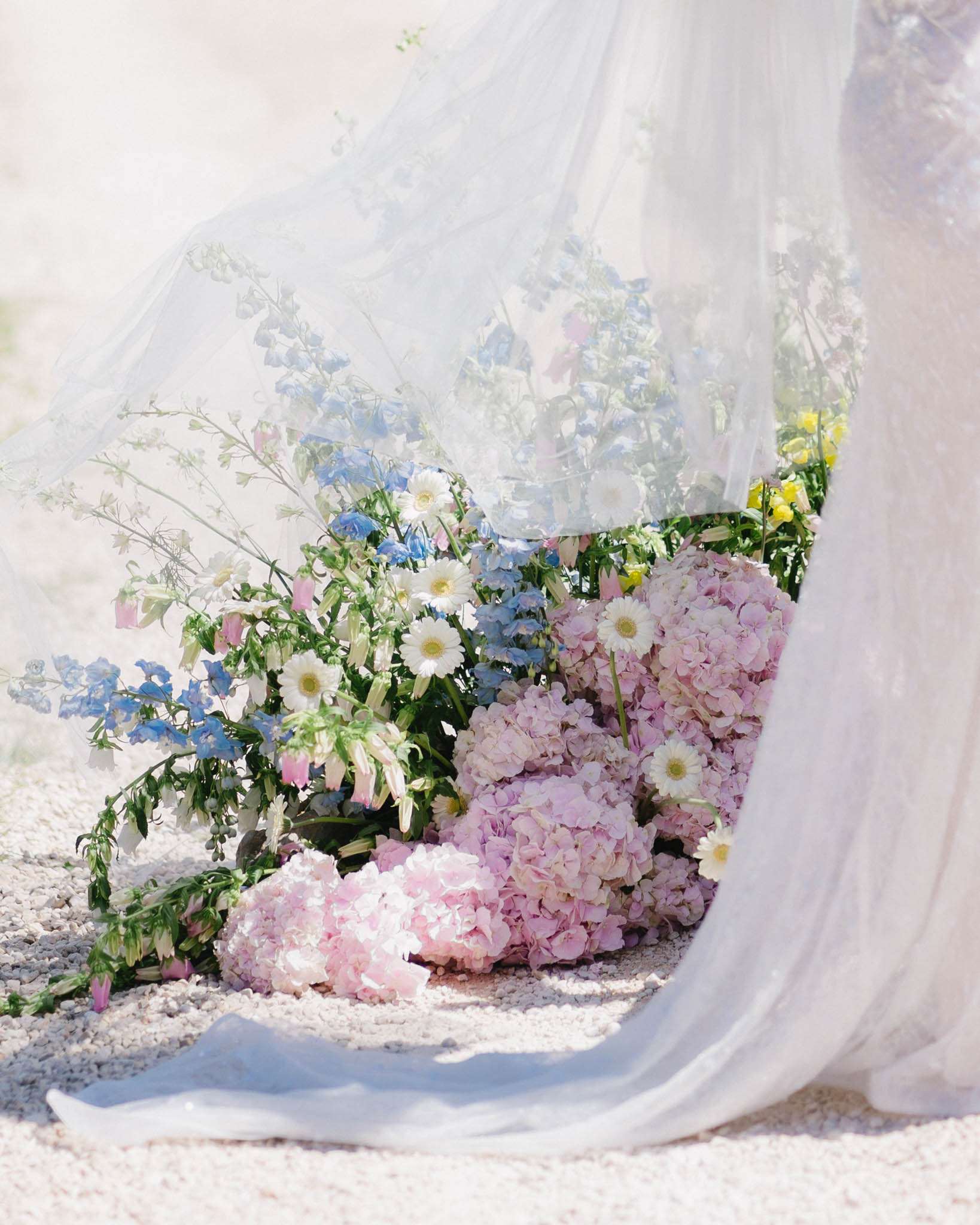 Pastel floral arrangement of pink hydrangeas, white daisies, and blue delphinium on white gravel with tulle overlay