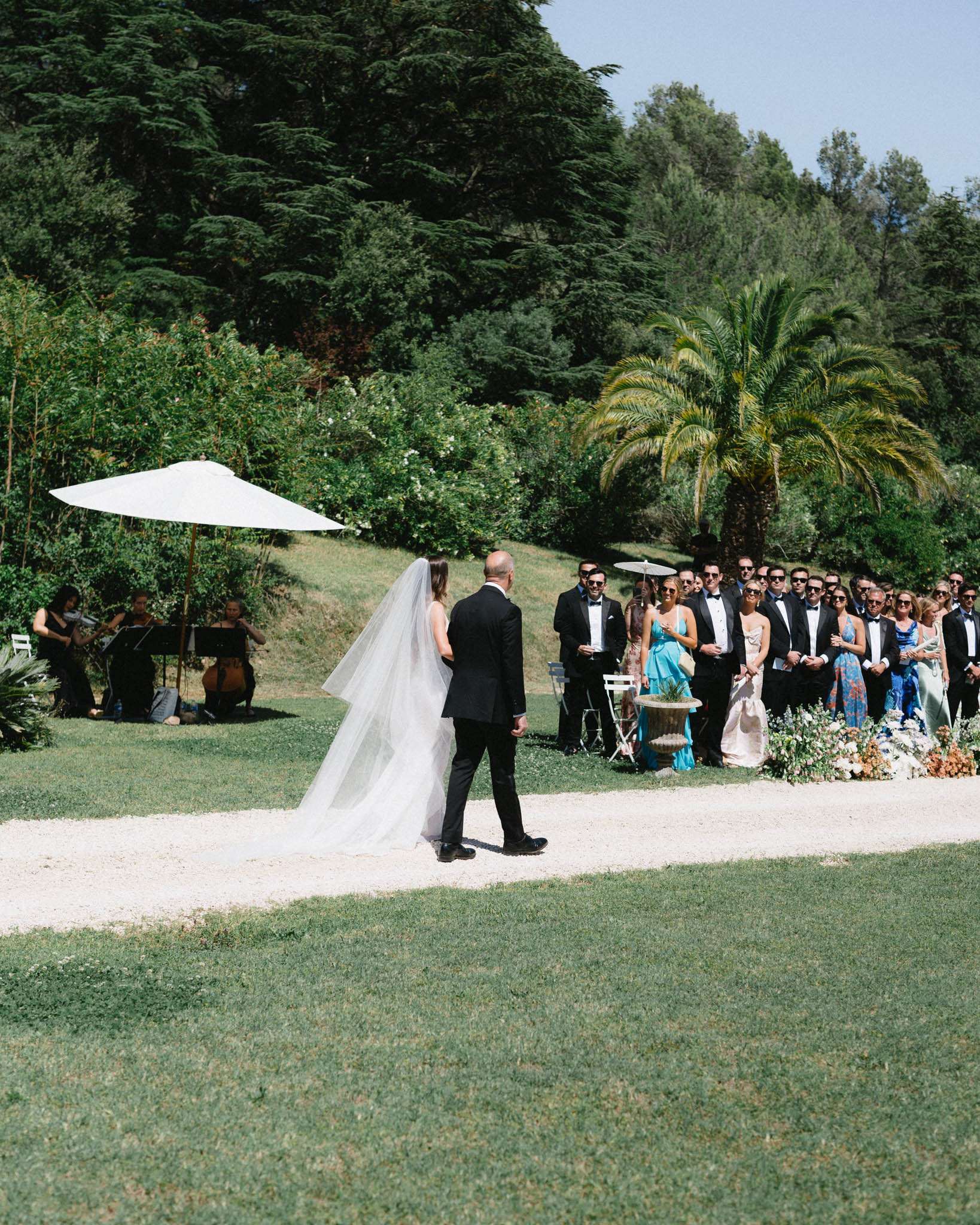 Outdoor garden ceremony with guests in semi-circle around couple and string trio under white umbrella