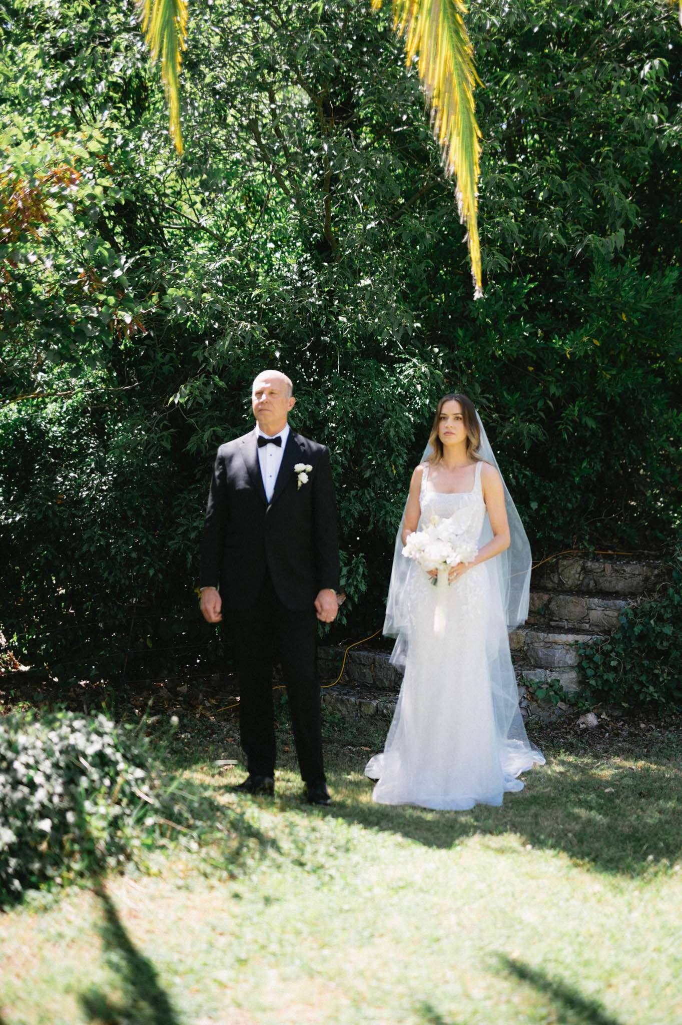 A bride and an older man, likely her father, stand side by side outdoors in a garden setting, appearing to wait before walking down the aisle. The bride wears a white fitted gown with a square neckline and delicate embellishment, paired with a long single-tier veil, and carries a bouquet of white flowers including what appear to be orchids and peonies. The man wears a classic black tuxedo with a black bow tie and a small white floral boutonnière on his lapel. The shot is a mid-distance portrait taken in natural daylight, with a low stone retaining wall visible in the background.