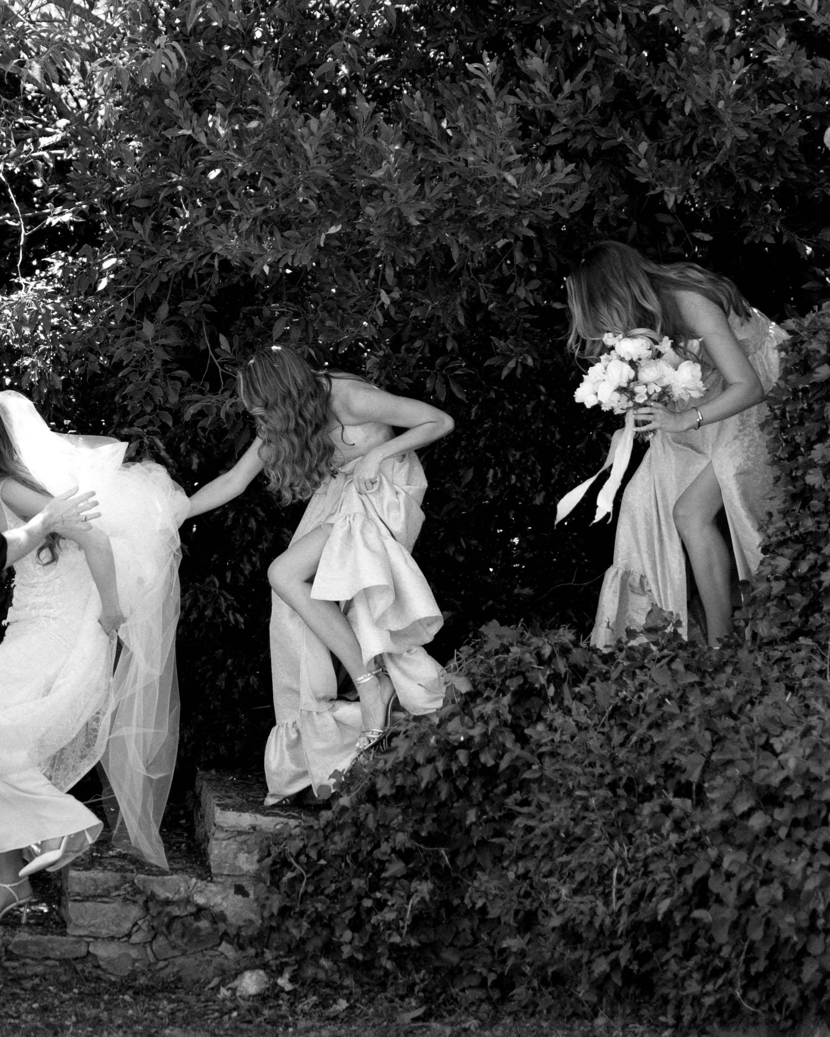 Black and white image of bride and two bridesmaids navigating stone garden steps holding bouquets