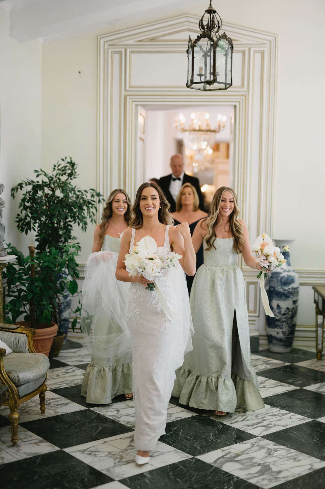 The bride is walking through an indoor hallway of what appears to be a classic French château or manor, heading toward the ceremony. She wears a fitted white gown with spaghetti straps, feather detailing, and a tulle overlay train, carrying a bouquet of white peonies and soft blush blooms. Two bridesmaids flank her in matching sage green jacquard full-length dresses with ruffled hems and square necklines, each holding a bouquet of blush, peach, and white flowers with trailing ivory ribbons. Behind the group, two adults — a man in a black tuxedo and a woman in a navy dress — follow through a doorway framed with white and gold molding, beneath which hangs a wrought-iron lantern with candles; a crystal chandelier is visible in the background room. The floor is a black and white marble checkerboard, and the interior décor includes gilded Louis XVI-style armchairs, blue and white chinoiserie porcelain vases, and potted greenery, reflecting a classic, formal French interior style. The shot is a medium portrait taken at eye level.