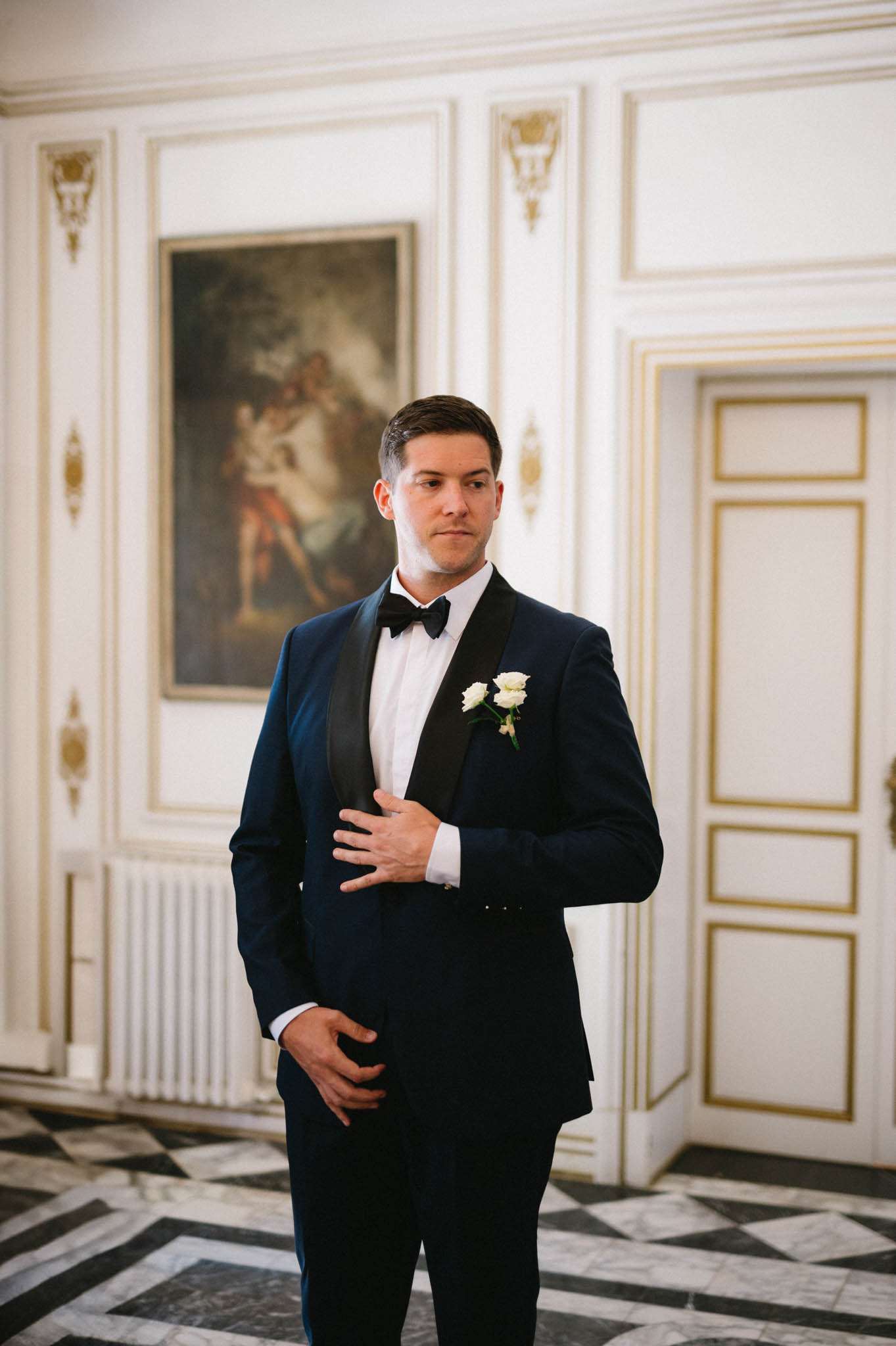 Groom in navy tuxedo with ivory rose boutonniere in ornate chateau room with boiserie and marble floor