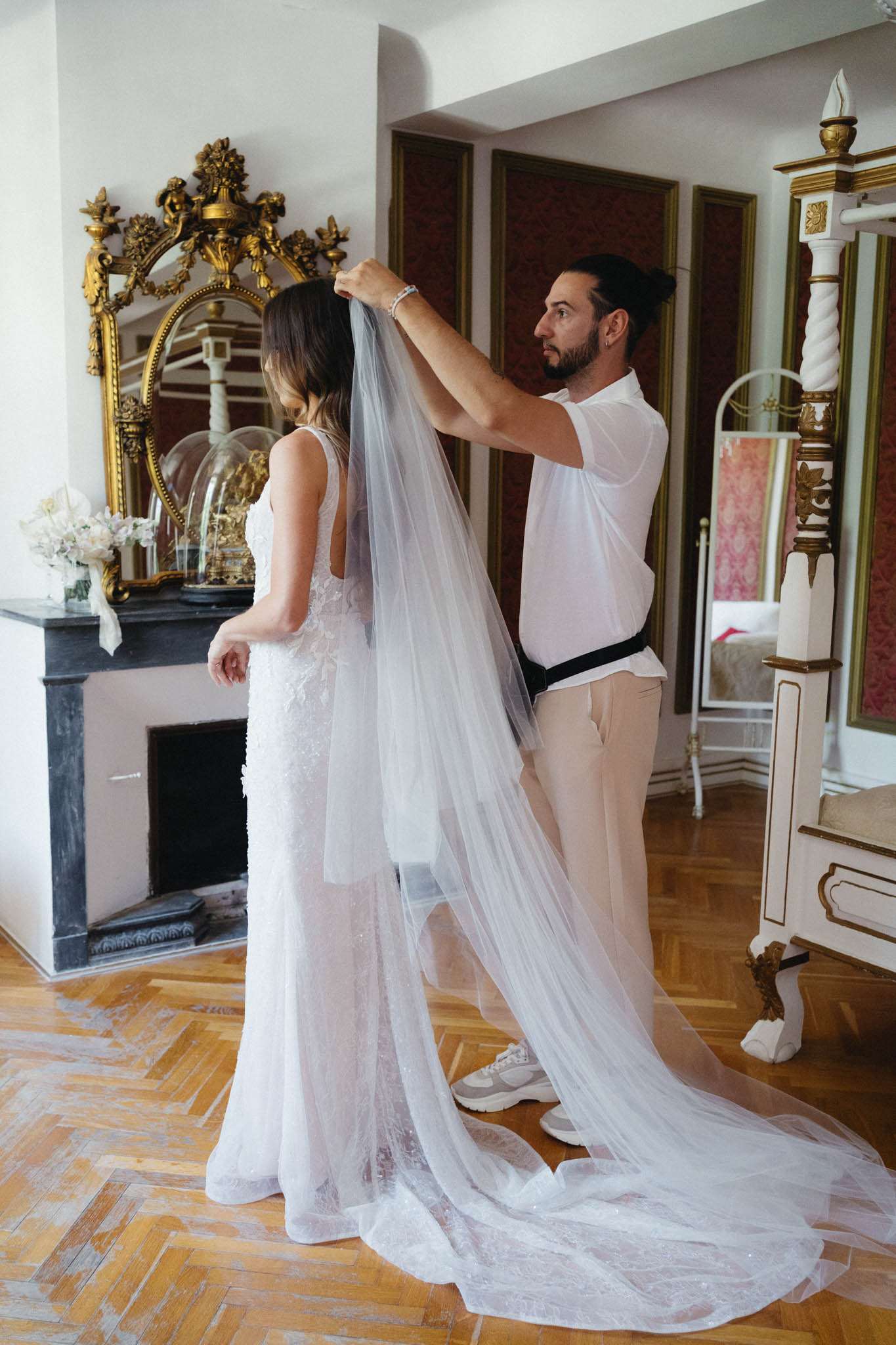 Stylist fastening cathedral veil to brides dark hair bride in low-back lace gown in red-panelled chateau room with fireplace