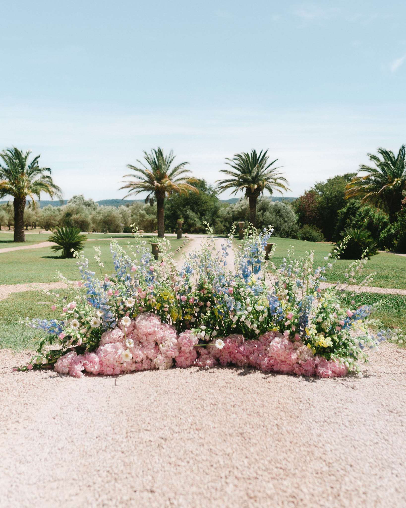 Ground-level floral installation with pink peonies and blue delphiniums on gravel garden path