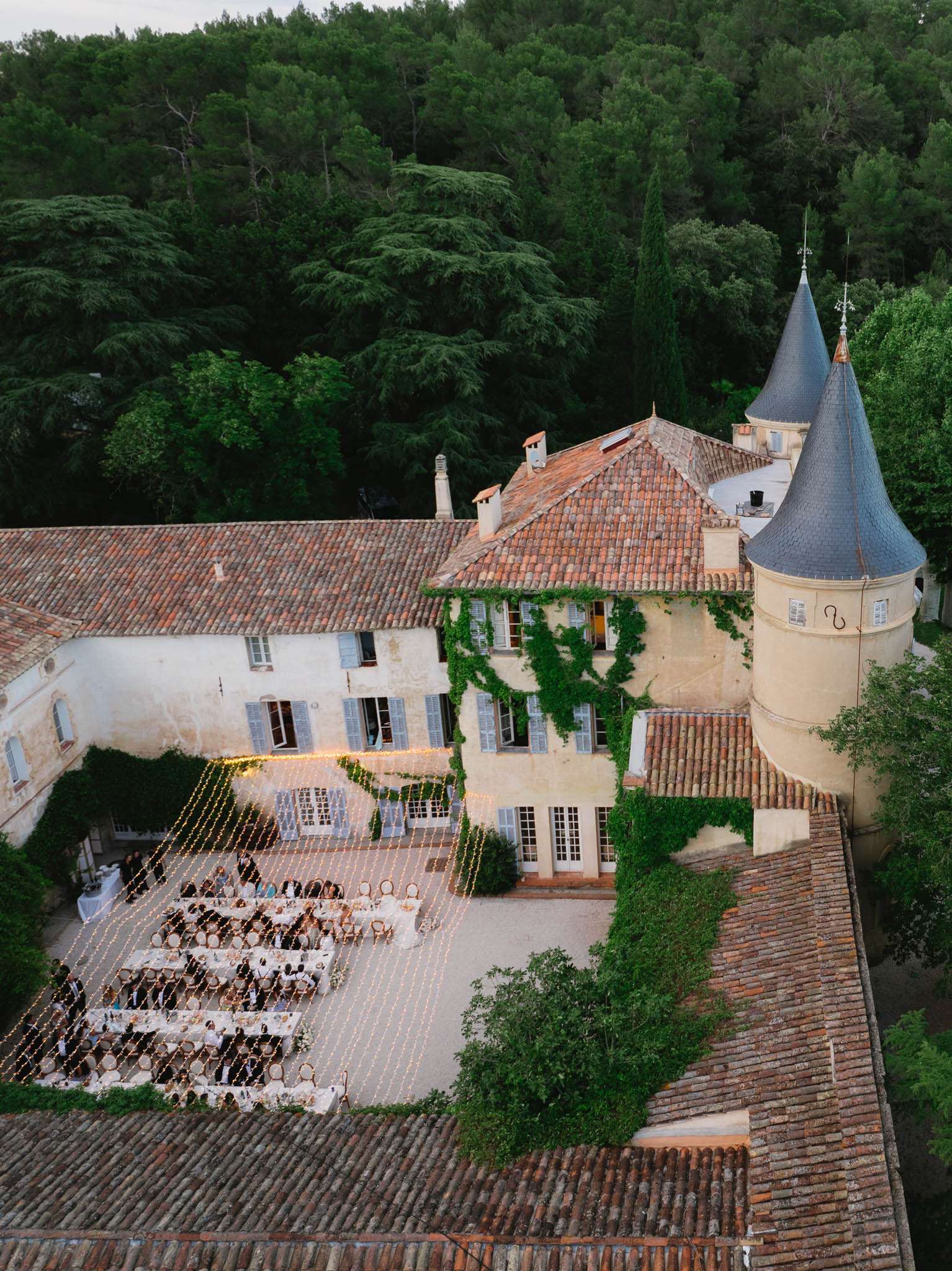 An aerial drone shot of an outdoor wedding reception dinner taking place in the courtyard of a French château, featuring terracotta-tiled roofs, blue-grey shutters, ivy-covered stone walls, and a distinctive conical turret with a slate roof. Guests — approximately 80 to 100 people — are seated at multiple long banquet-style tables arranged in a rectangular formation across the gravel courtyard. The tables are dressed in white linens and set with what appears to be glassware and centerpieces. A grid of warm golden fairy lights is strung low across the entire dining area at rooftop level, creating a lit canopy effect over the guests as dusk settles. The surrounding buildings form a U-shaped enclosure around the reception space, and dense woodland frames the estate from behind. Potential venue feature image.