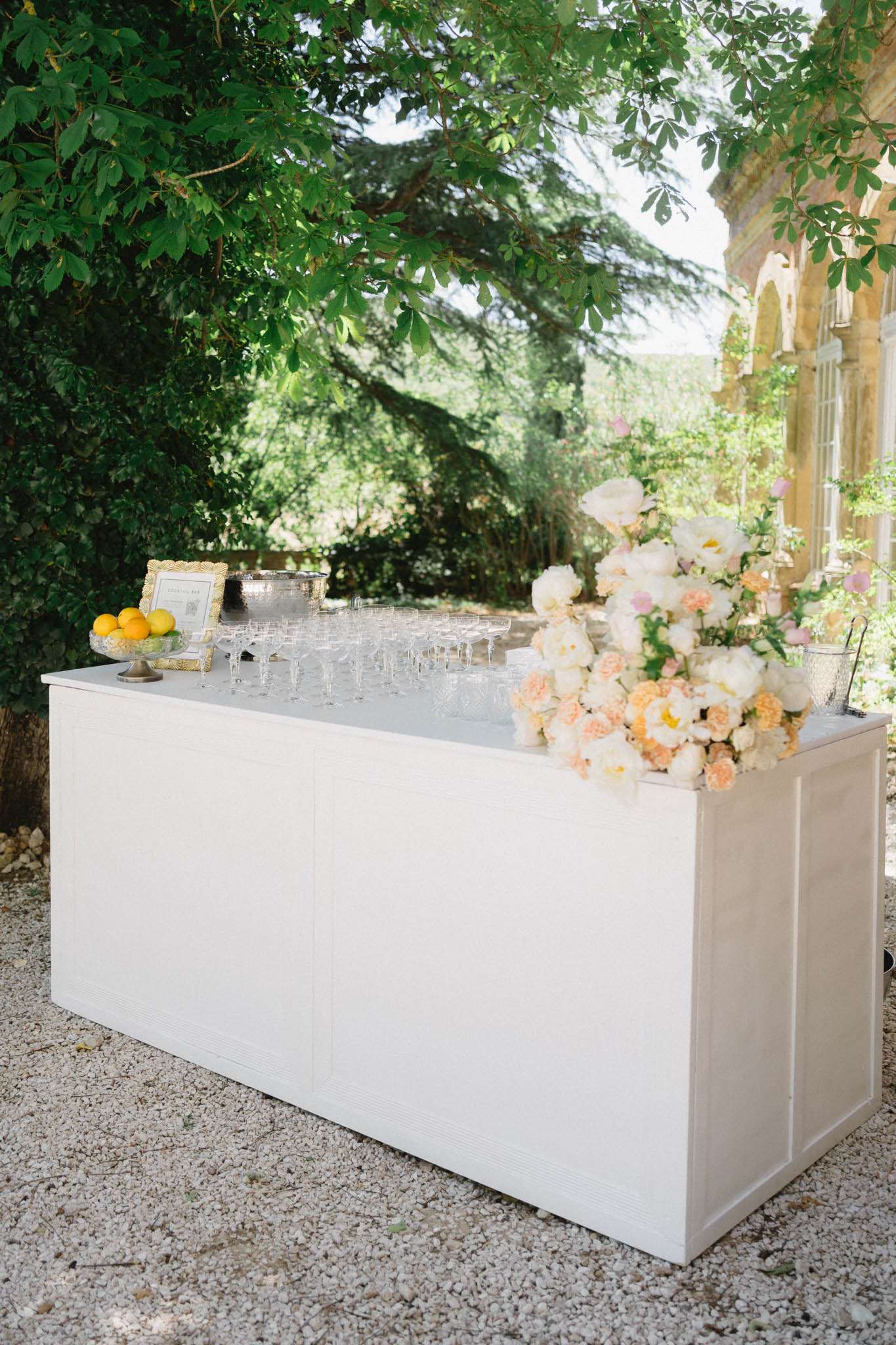 White cocktail bar with peony and peach rose arrangement, coupe glasses, and gold-framed menu on terrace