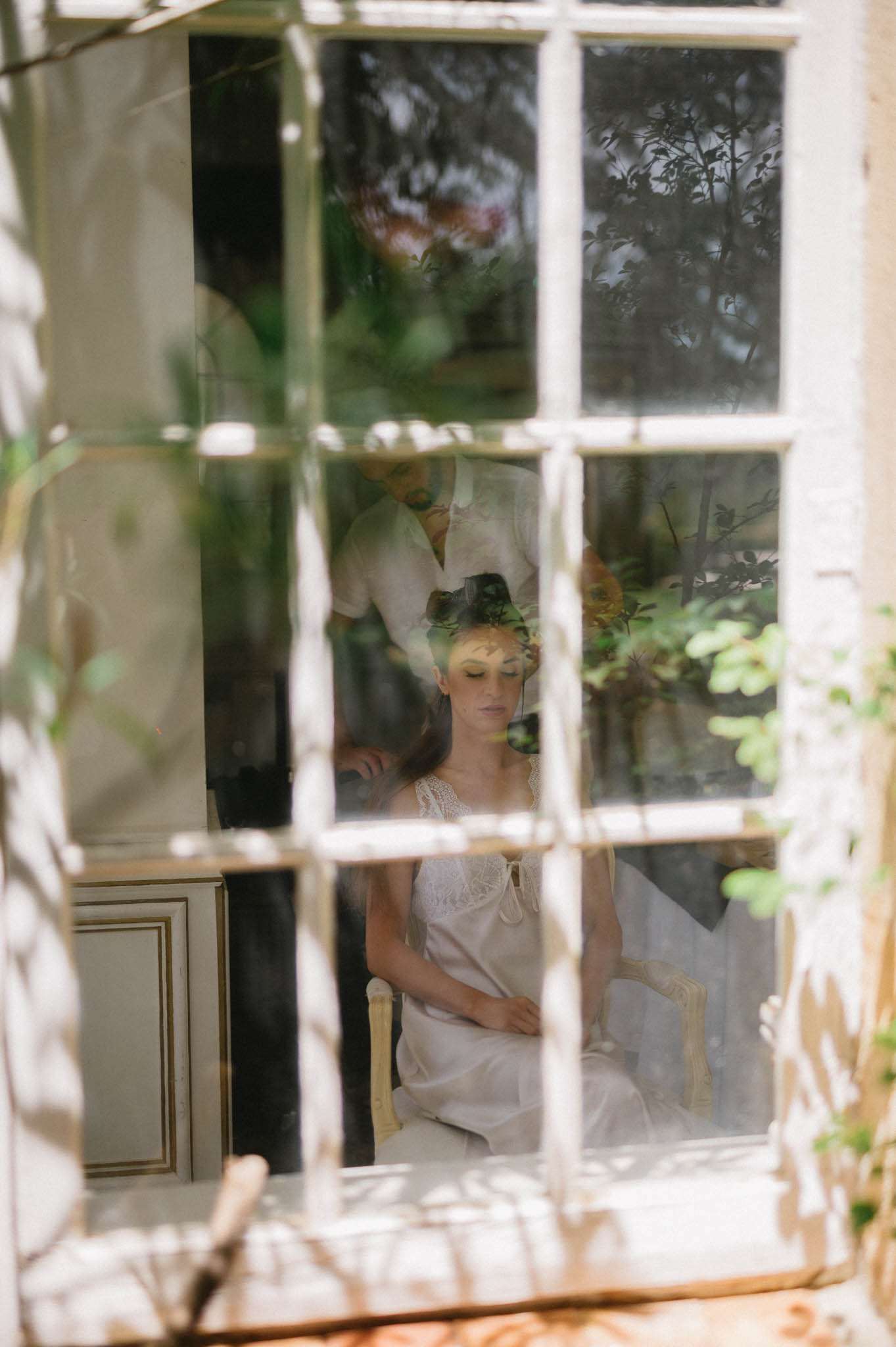 Bride seated inside chateau room while stylist places floral crown on her head, viewed through French window