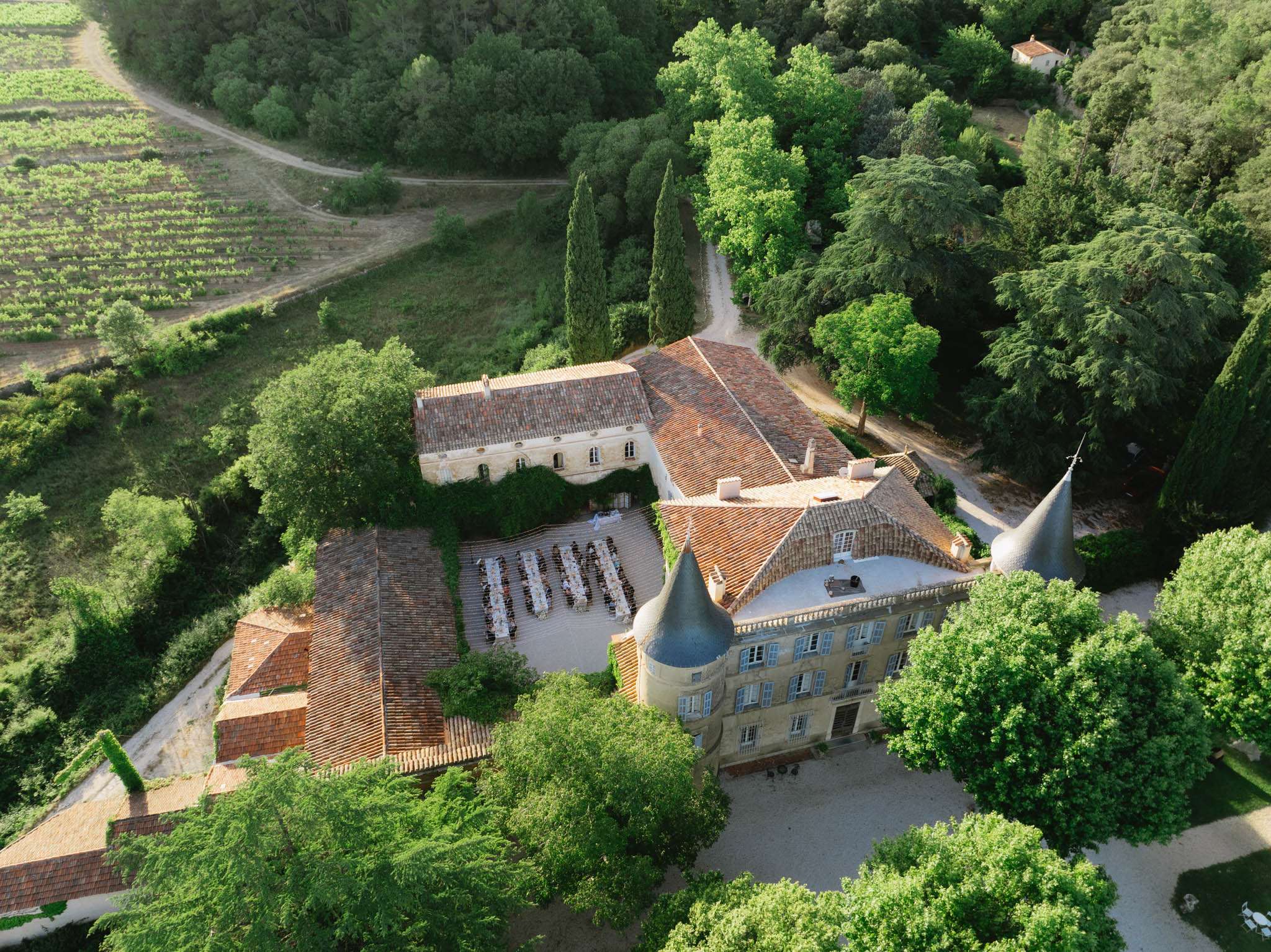 Aerial drone view of chateau courtyard wedding reception with long white dining tables, terracotta roofs, and surrounding ...
