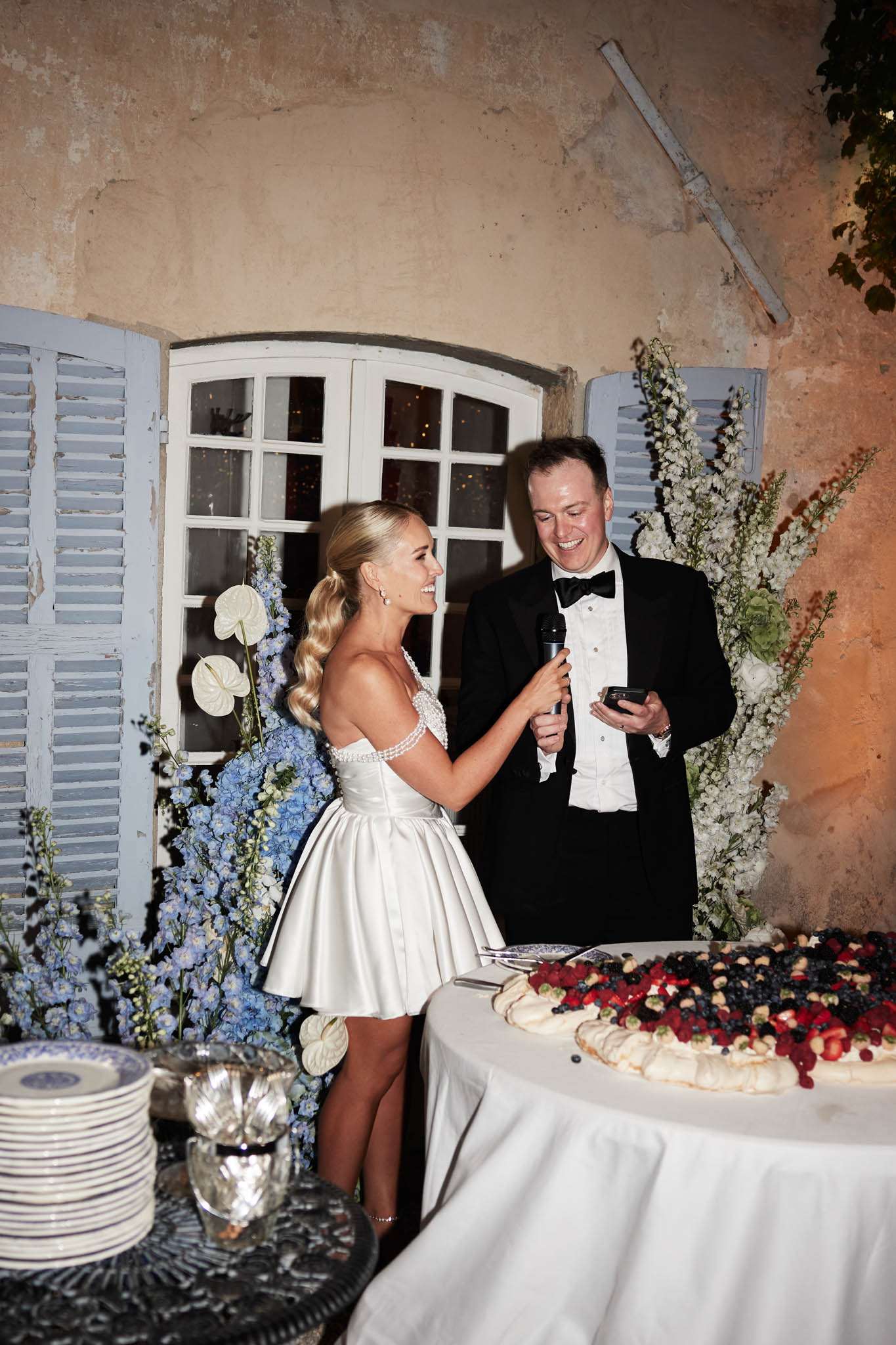 Bride and groom laughing during speech beside pavlova cake with blue delphinium arrangements at stone wall