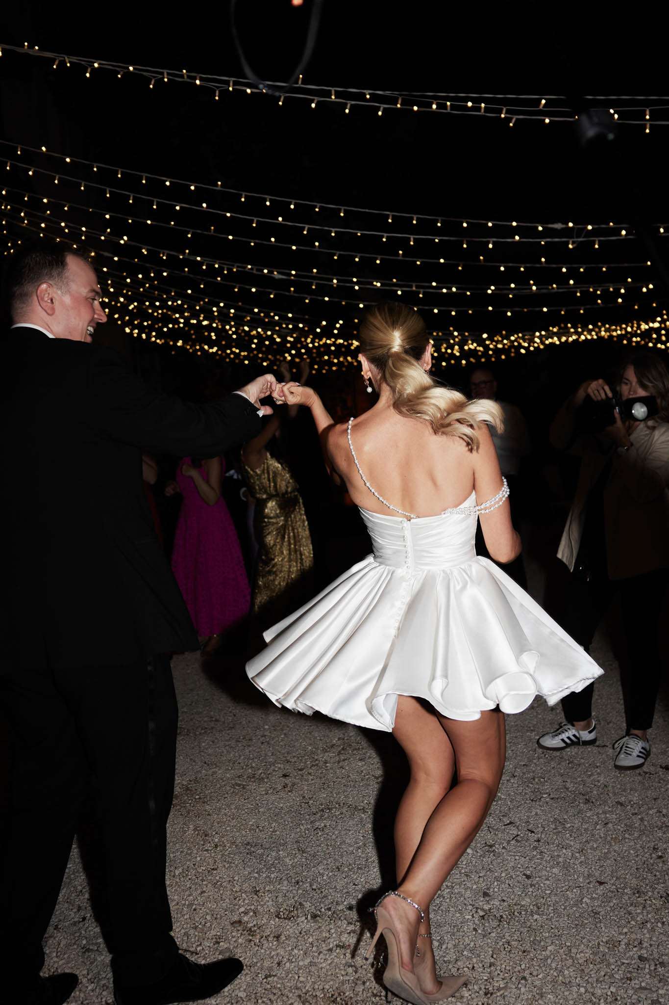 Bride in short white reception dress spinning on outdoor dance floor under fairy light canopy at night