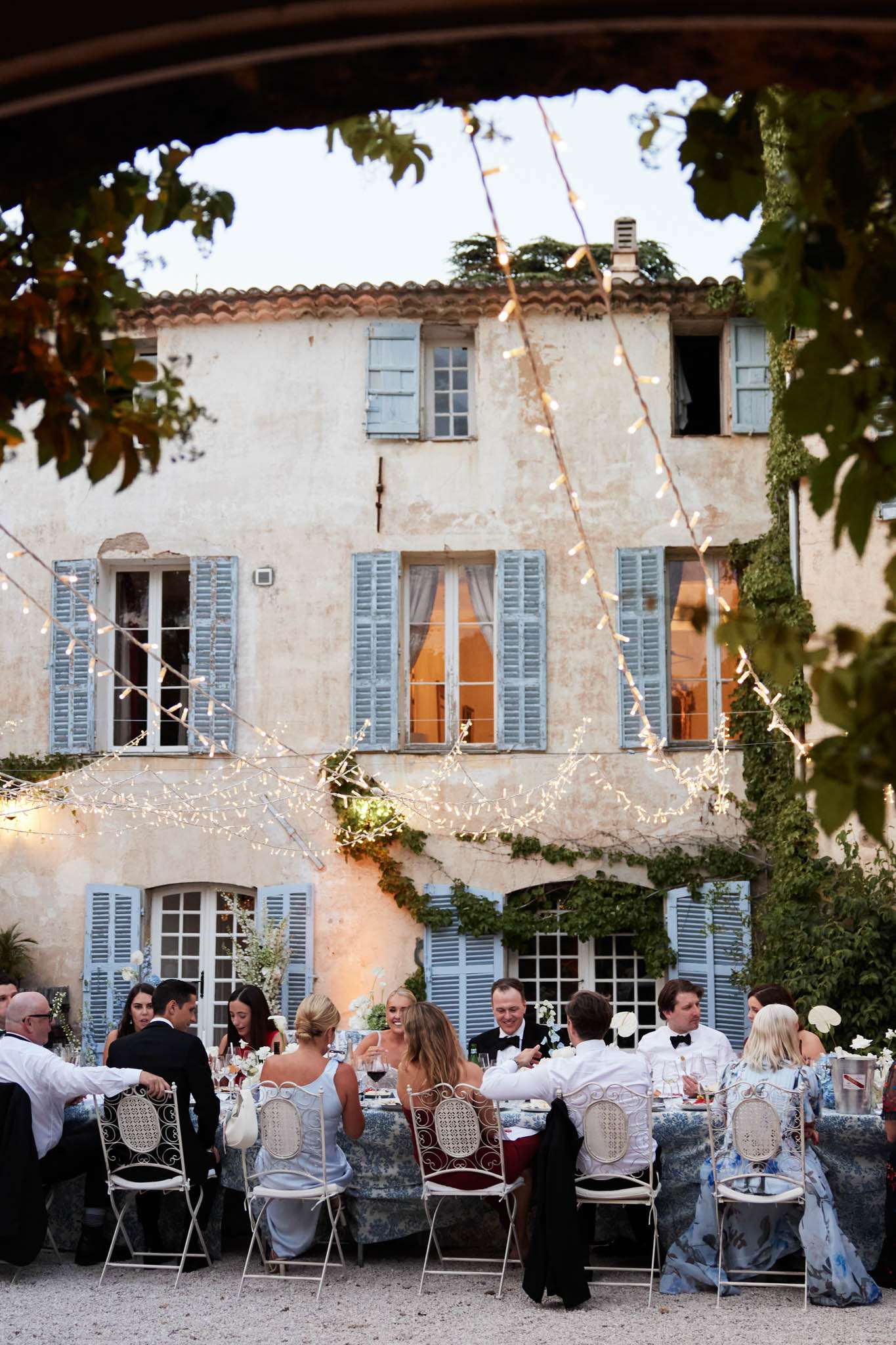 Dusk reception in Provencal courtyard with fairy lights, toile tablecloths, and wrought-iron chairs