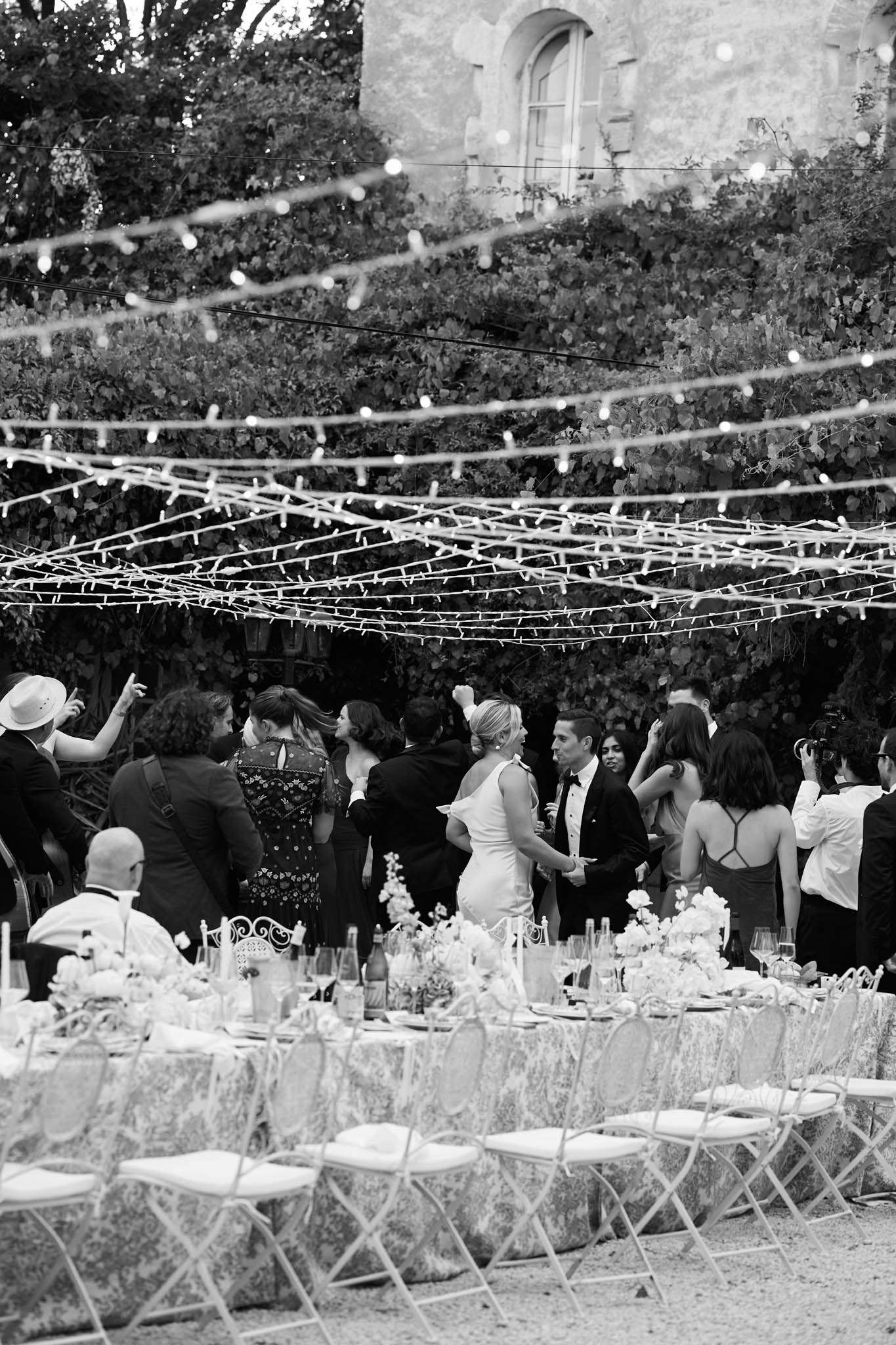 A black-and-white wide shot of an outdoor wedding reception in the grounds of what appears to be a French château or manor, with ivy-covered stone walls visible in the background. Multiple strings of globe fairy lights and dense rows of fairy lights are draped overhead in a canopy formation, creating strong contrast against the dark foliage behind. The bride, wearing a fitted sleeveless gown, stands near the groom in a tuxedo among approximately 15–20 mingling guests. Long reception tables are set with patterned tablecloths, floral centerpieces, wine bottles, glassware, and place settings, with ornate white metal chairs and folding bistro-style chairs arranged around them. The overall styling reads as classic French outdoor reception with a formal table setup and festive overhead lighting.