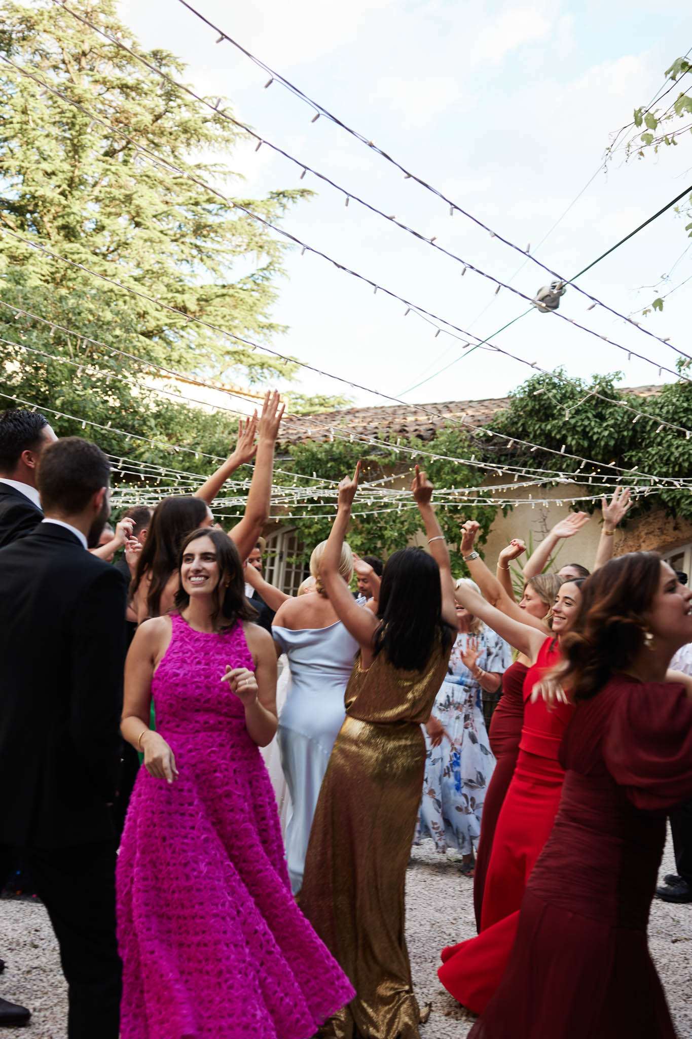 Twenty guests dance with arms raised under fairy lights in stone courtyard with ivy-covered walls
